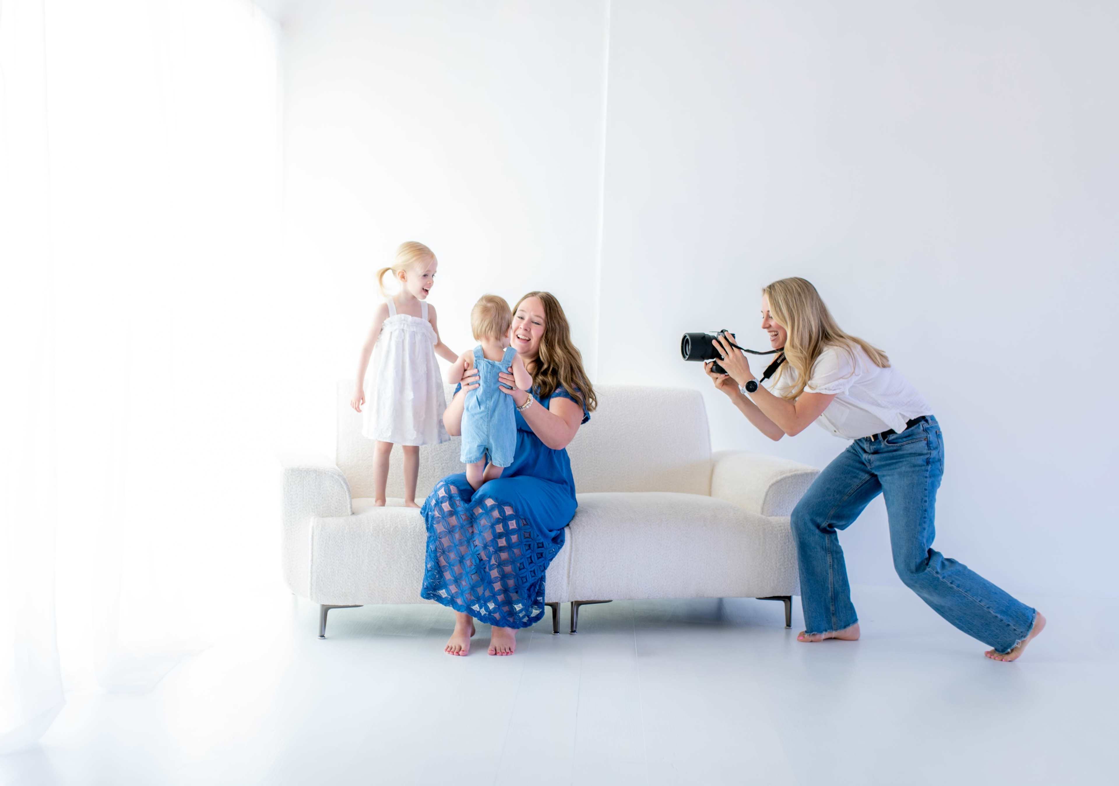A woman takes photos of a mother and her two young children on a couch in a brightly lit room.