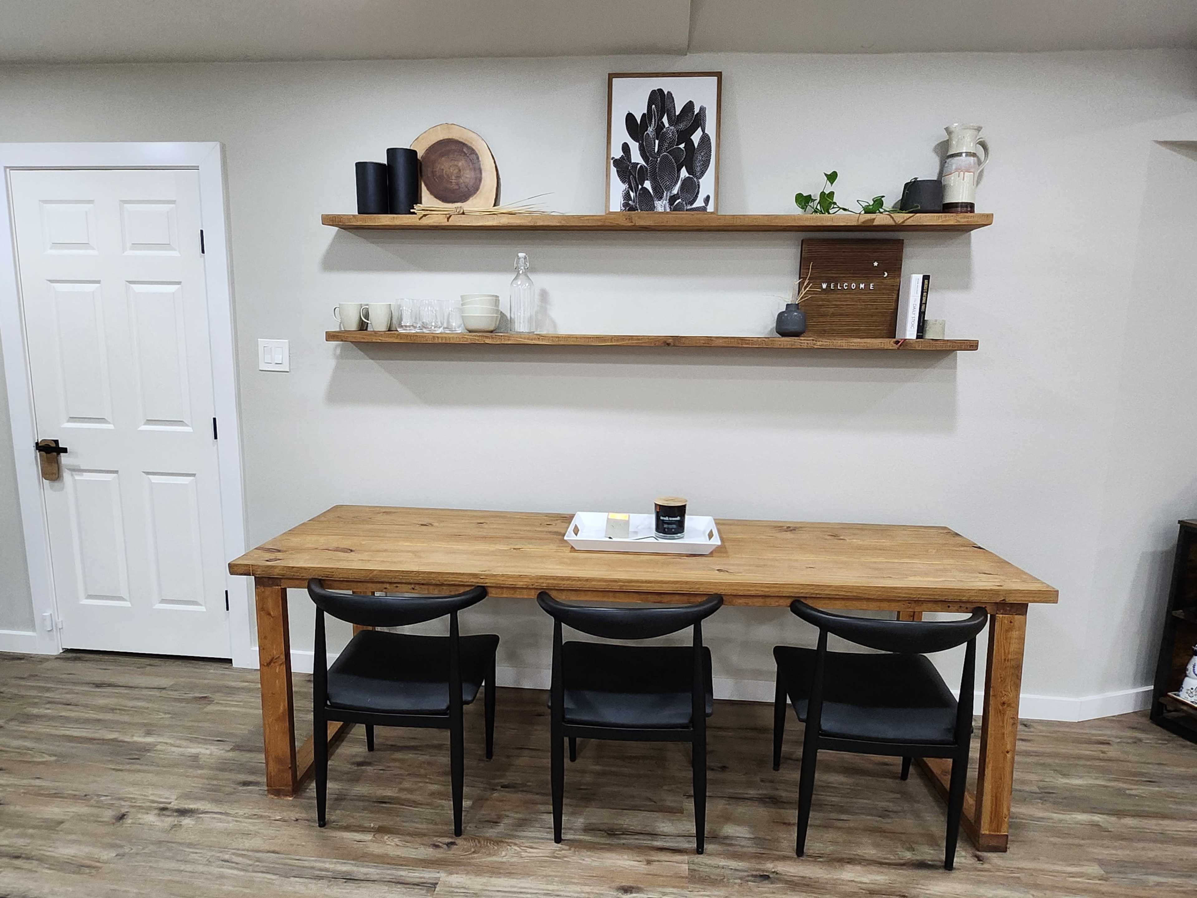 A wooden dining table with three black chairs is positioned under two shelves filled with decorative items, against a light-colored wall.