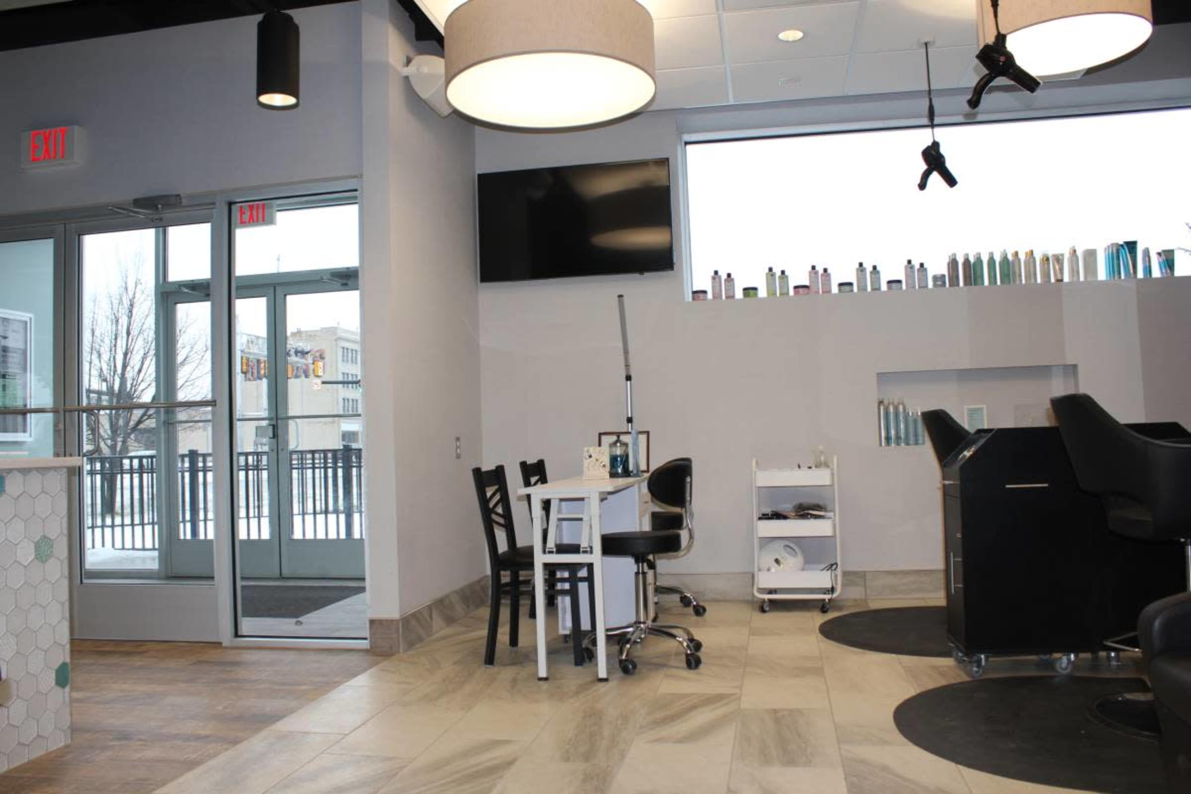 The image shows a modern salon interior with a reception area, a treatment table, and products displayed on shelves near large windows.