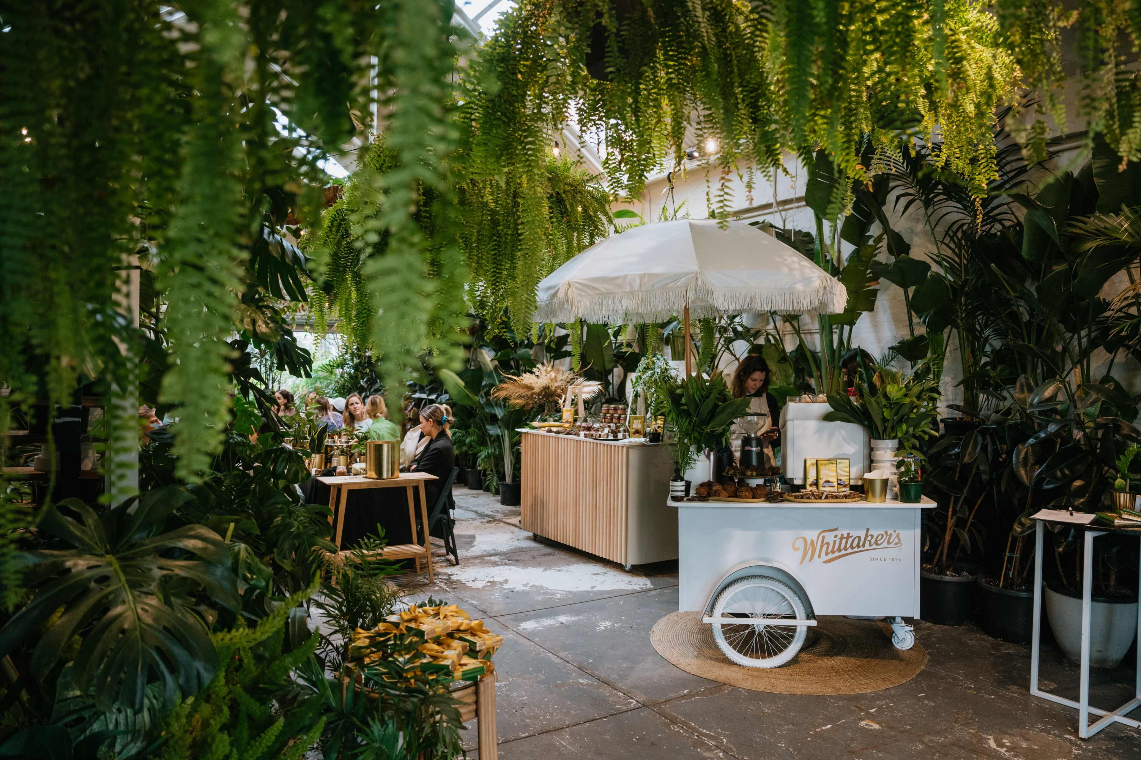 A cozy indoor space filled with lush green plants, featuring a small service cart under an umbrella and people seated at tables in the background.