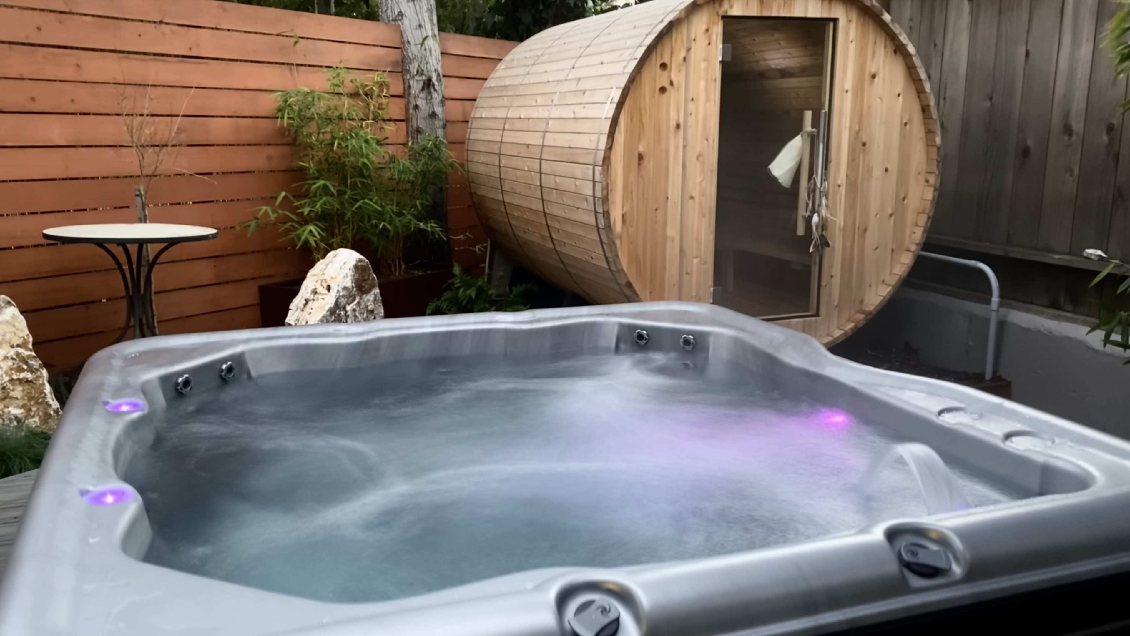 A hot tub with colored lights is positioned in front of a wooden sauna and surrounded by plants.