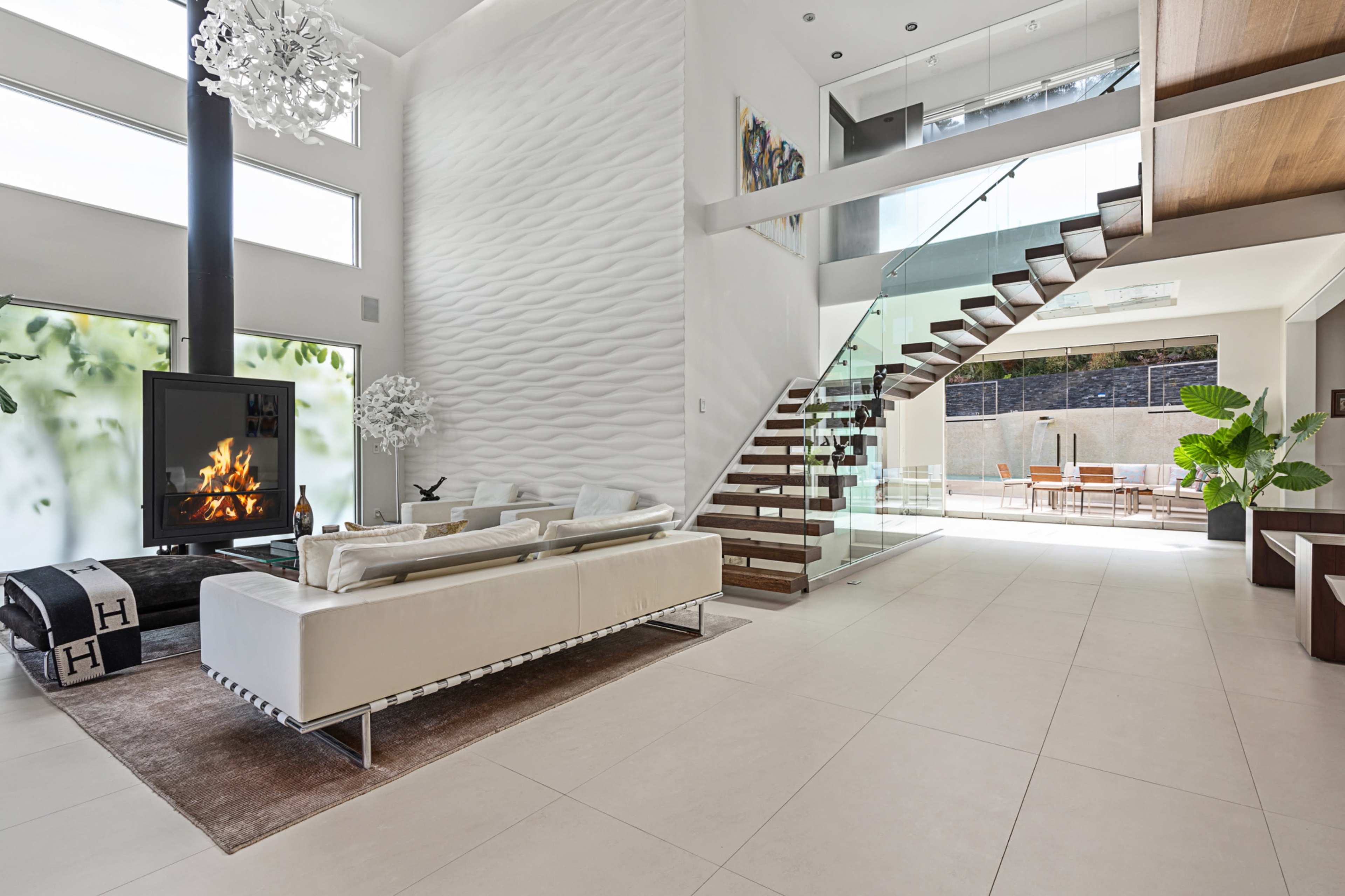 The image shows a modern living room with a tall ceiling, featuring a white sectional sofa, a fireplace, large windows, and a staircase with wooden steps.