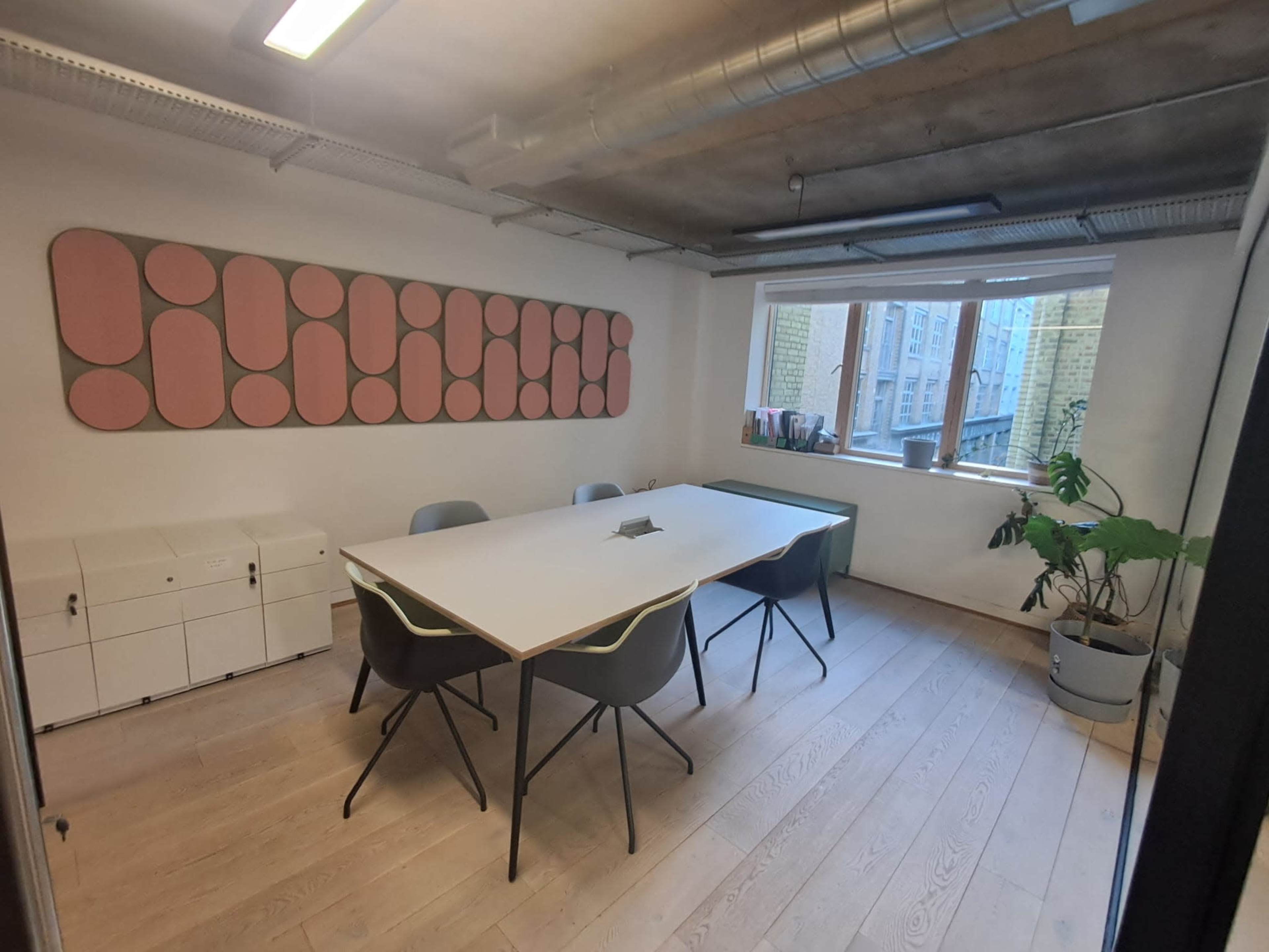 The image shows a modern meeting room with a large white table, several chairs, and a window featuring a green plant and a colorful wall decoration.
