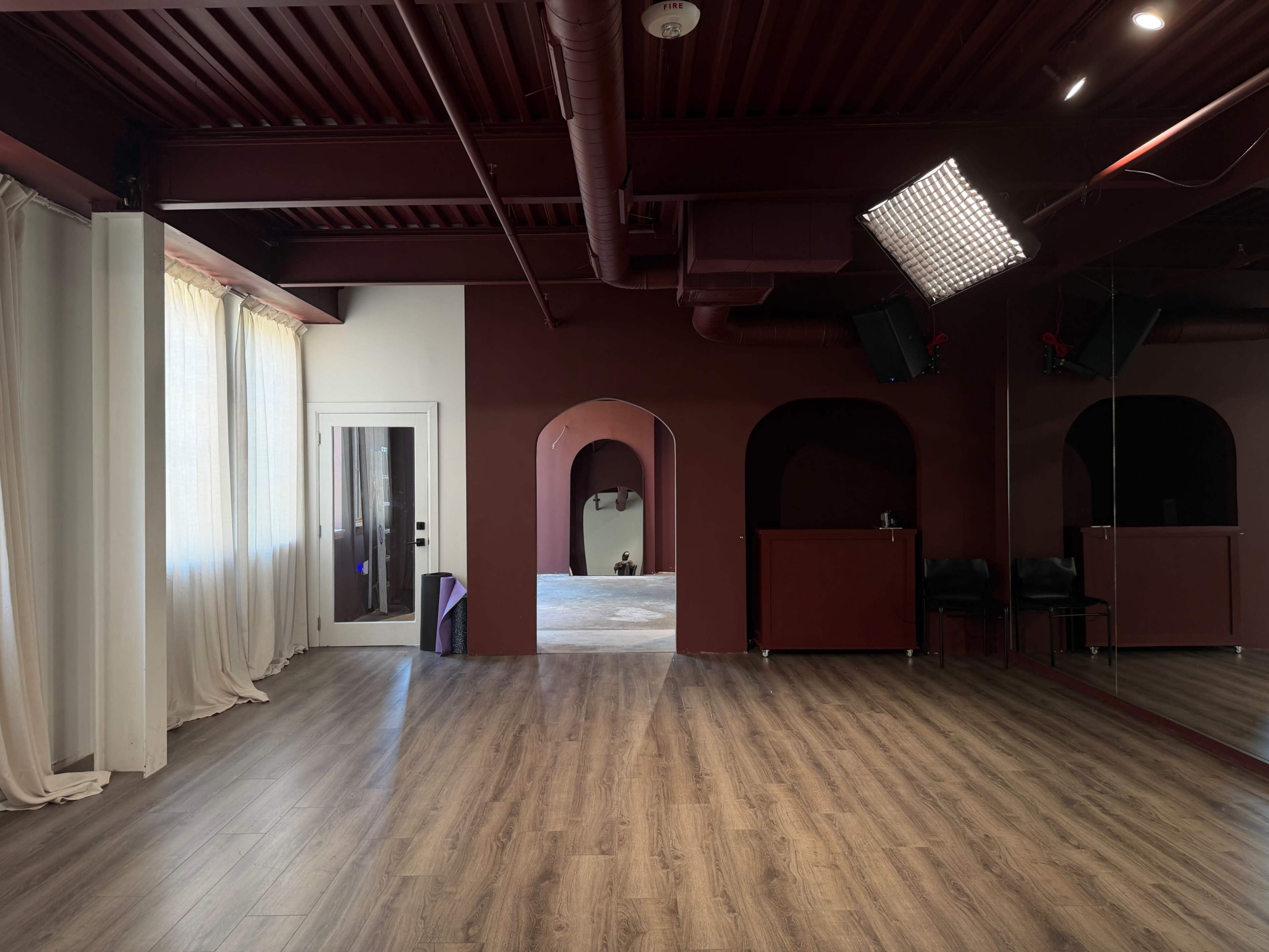 The image shows a spacious room with wooden flooring, red walls, large windows covered with white curtains, and a mirror on one side.
