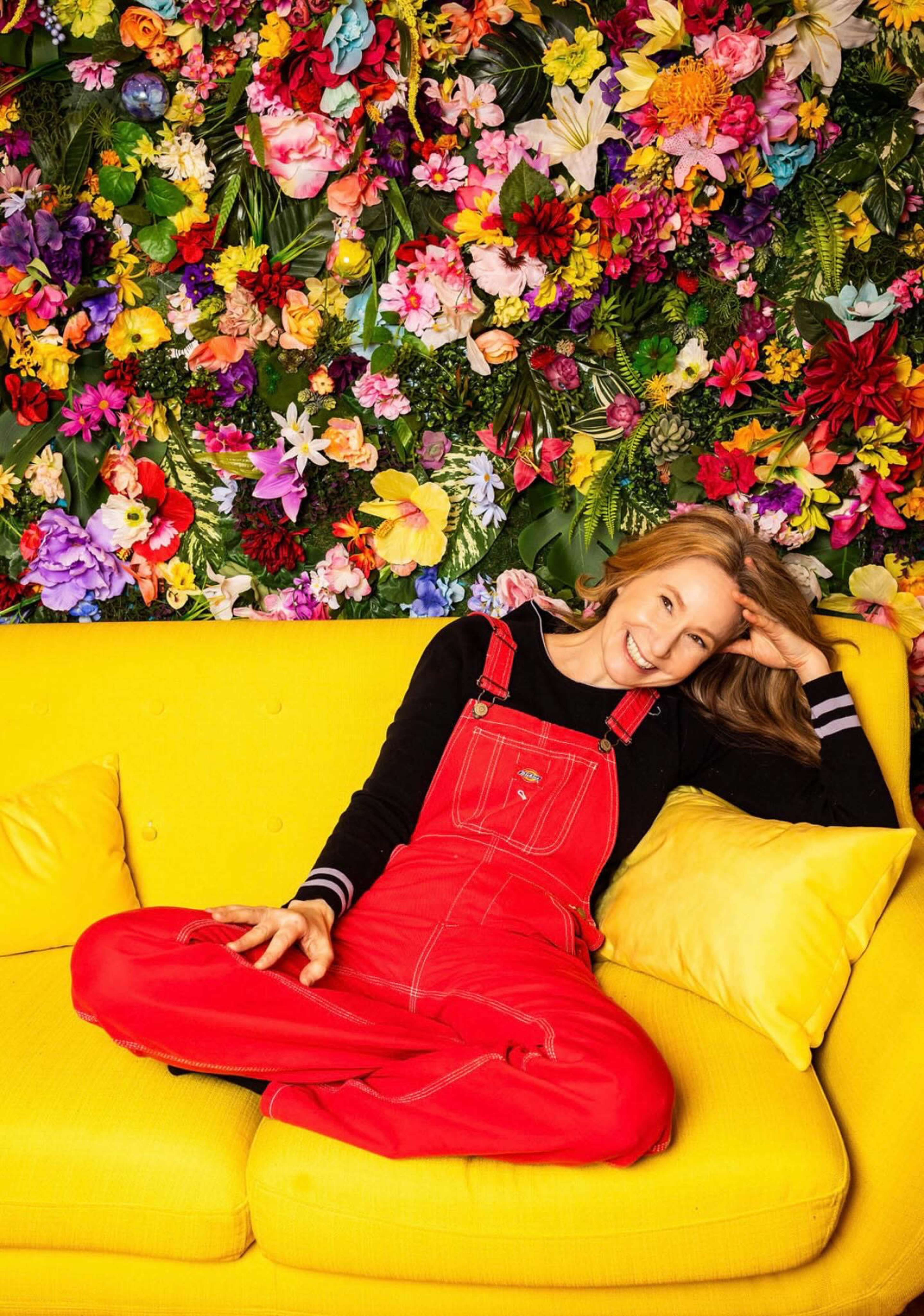 A woman in red overalls sits on a yellow couch against a vibrant floral backdrop.