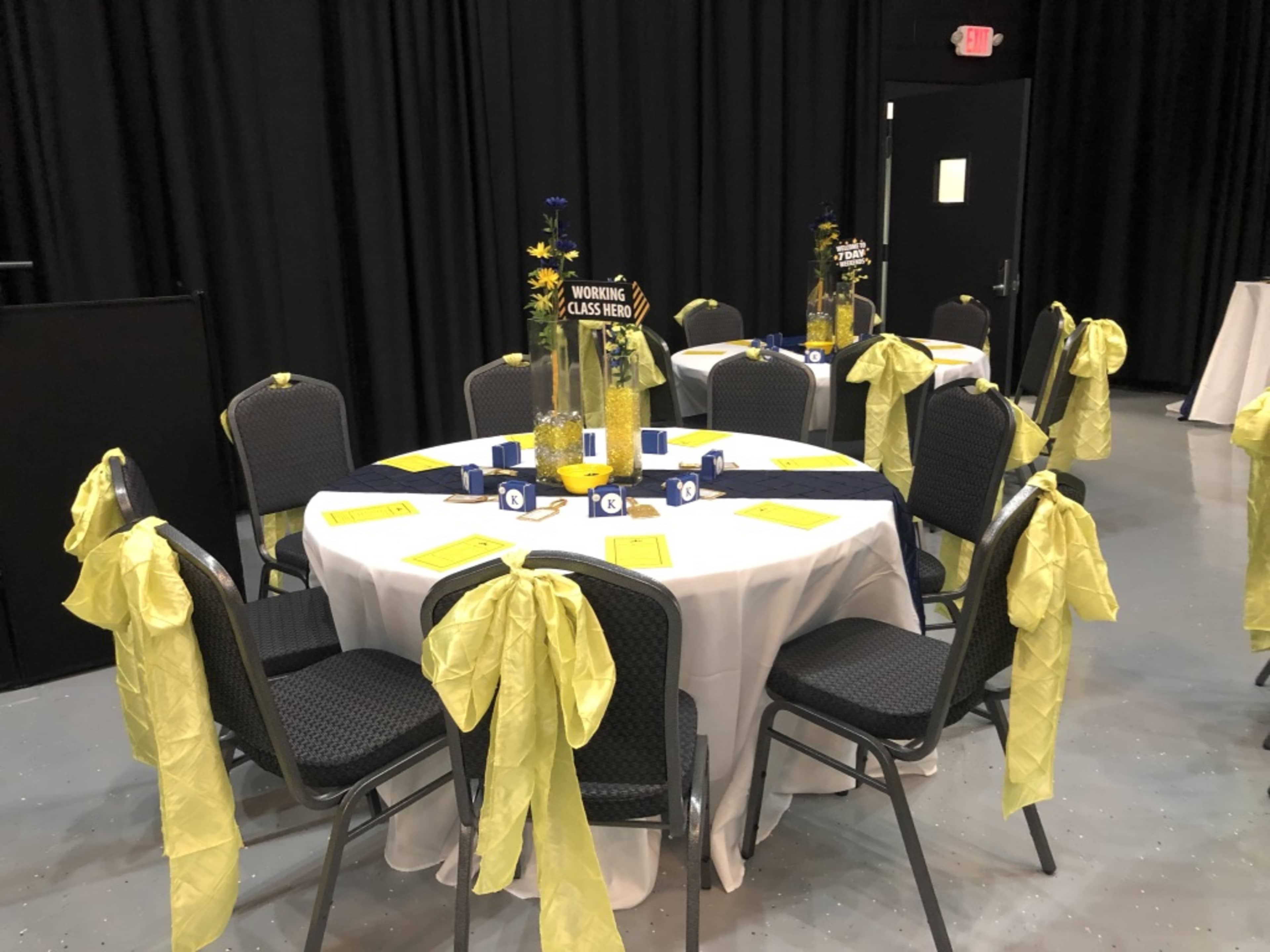 A round table is set with yellow and blue decorations, featuring place cards and tall centerpieces, in a dimly lit venue.