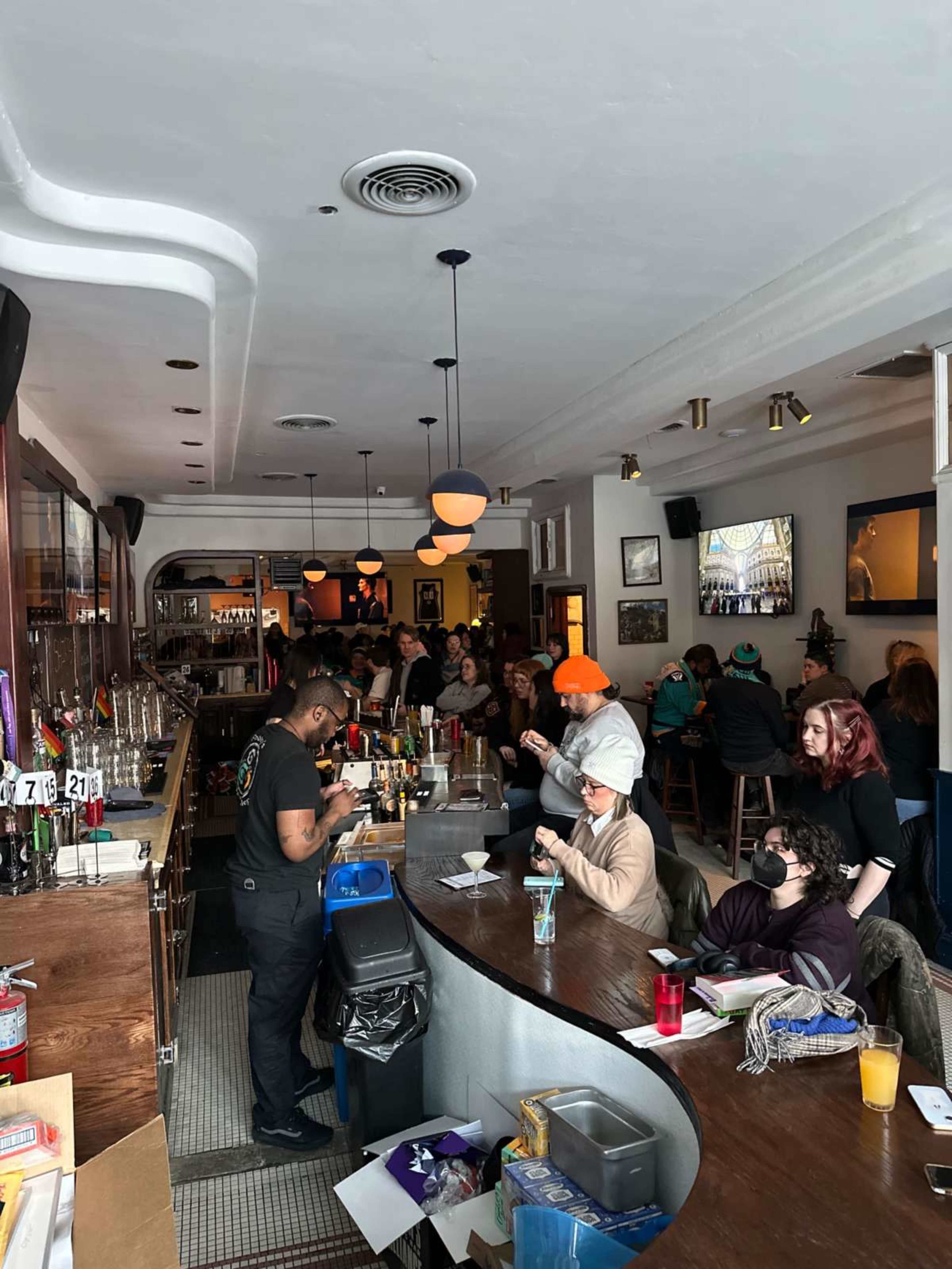 The image shows a lively bar interior with patrons seated at tables and a long curved bar, where a bartender is preparing drinks.