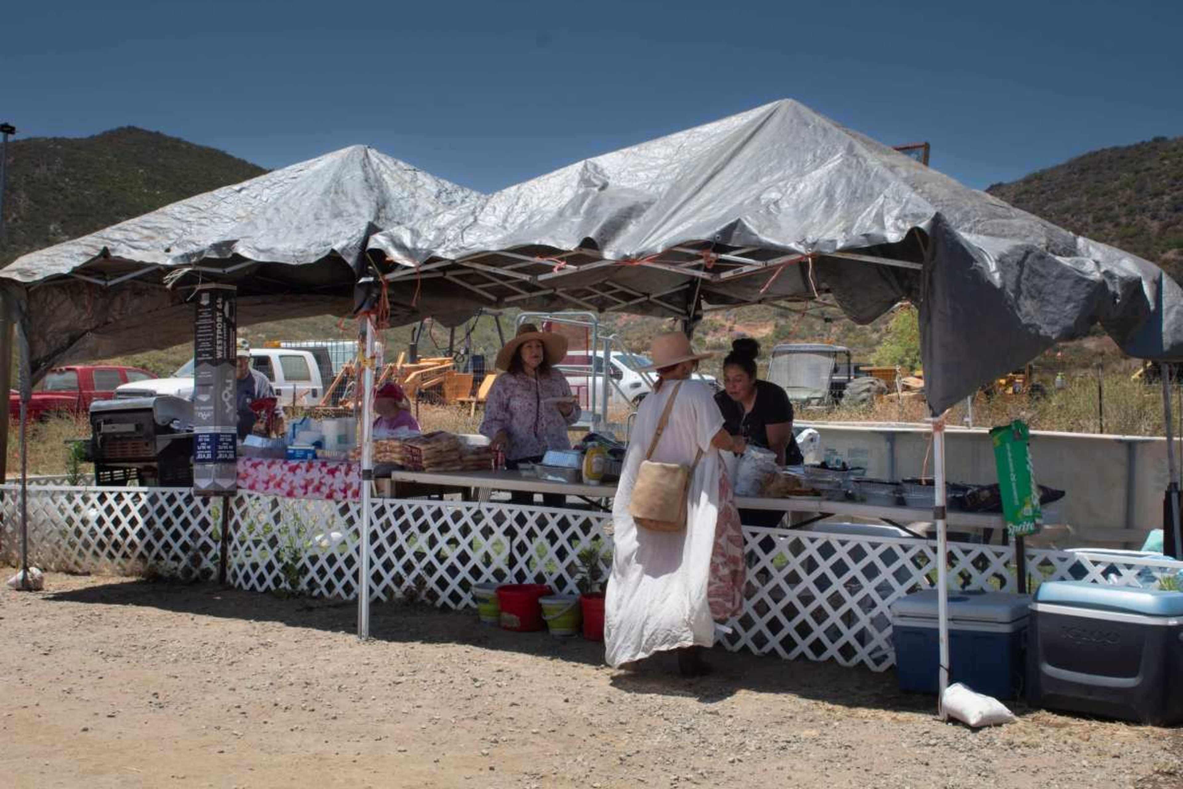 A market tent with vendors selling goods is set up in an outdoor area, surrounded by vehicles and hills.