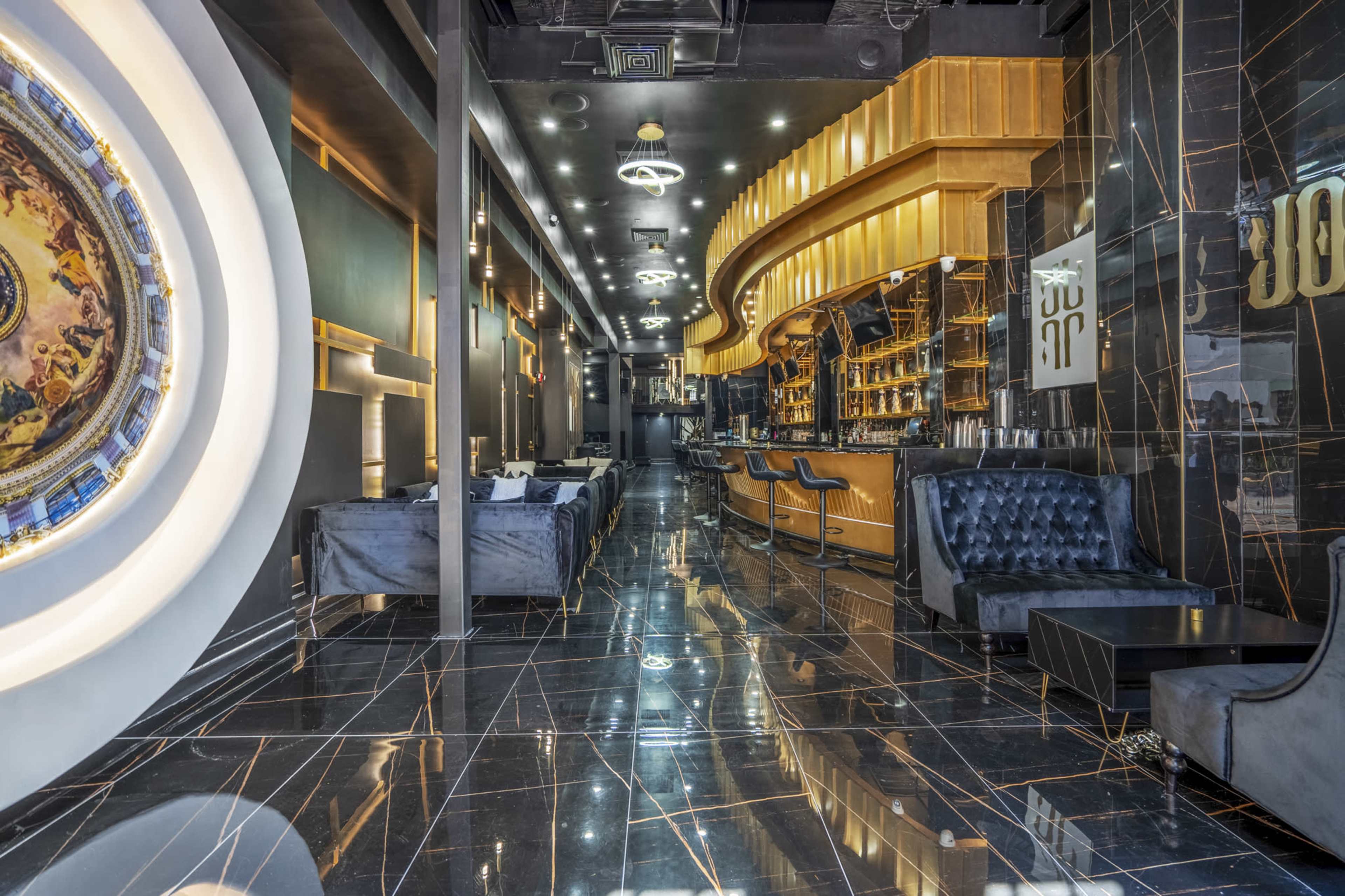 A modern bar interior with a polished black marble floor, elegant seating areas, and a long, illuminated bar counter in the background.