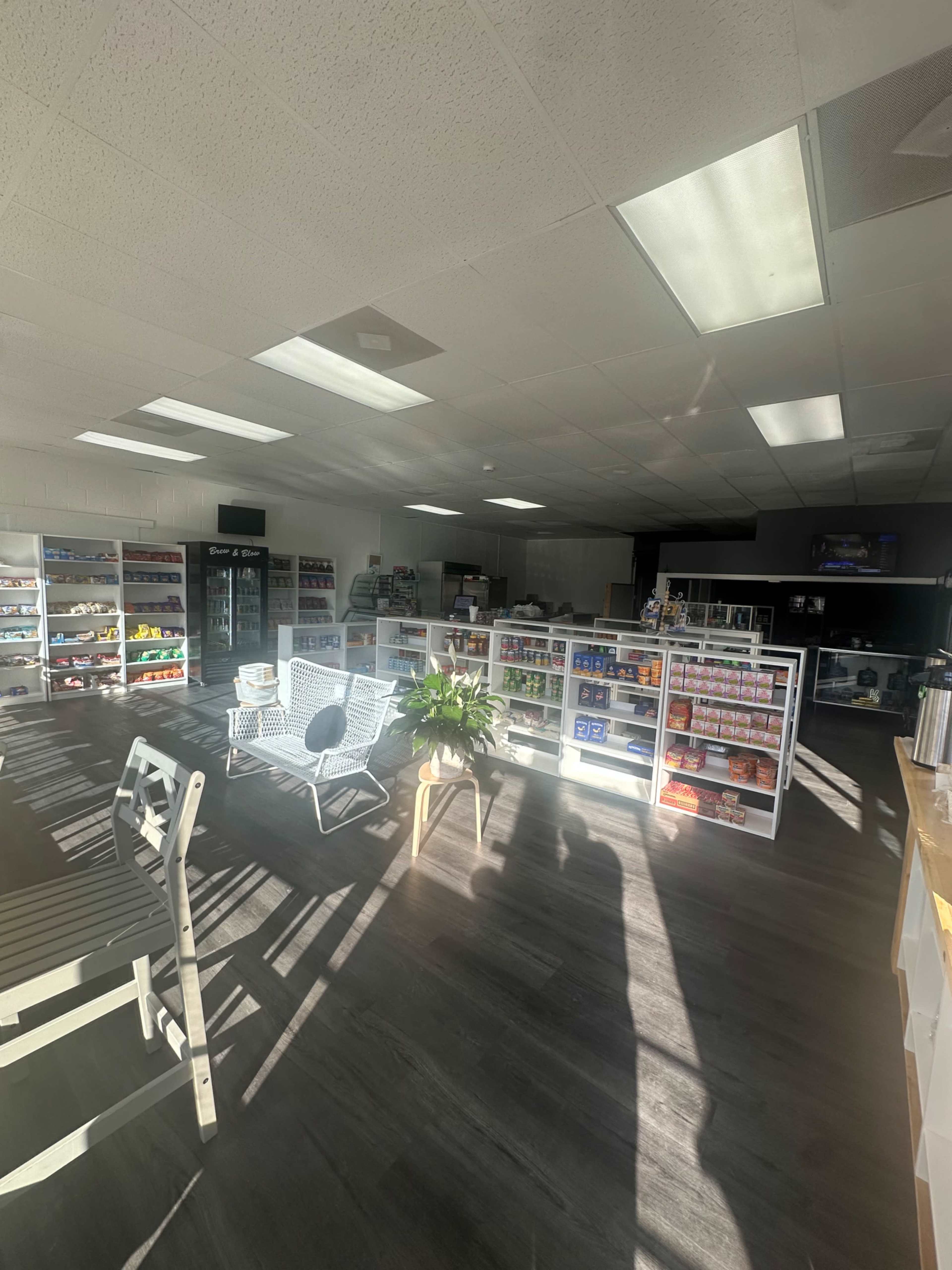 The image shows a brightly lit convenience store interior with shelves displaying various products and a seating area with a chair and potted plant.