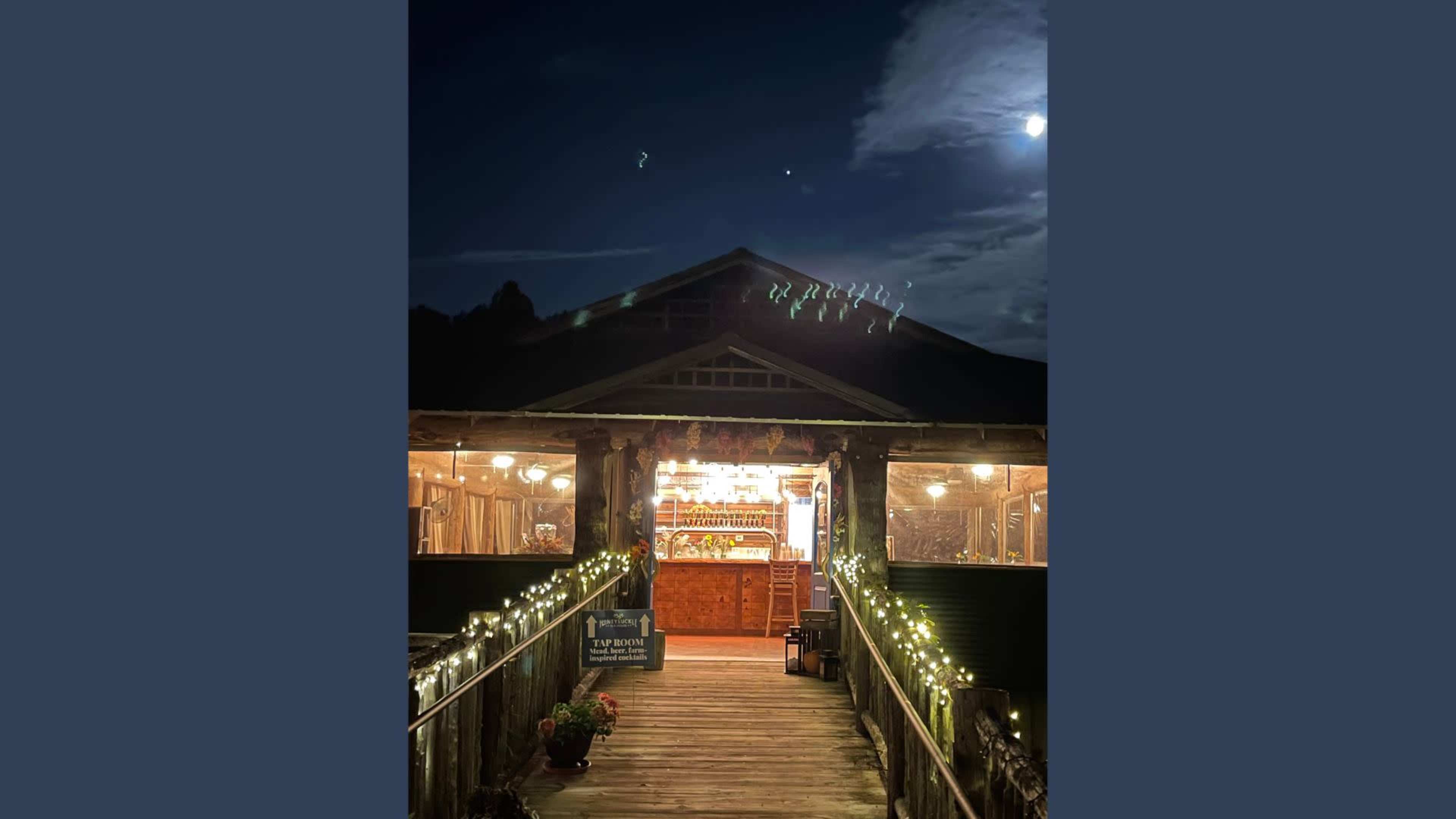 A wooden building with large windows is illuminated at night, connected by a lit pathway and surrounded by greenery.