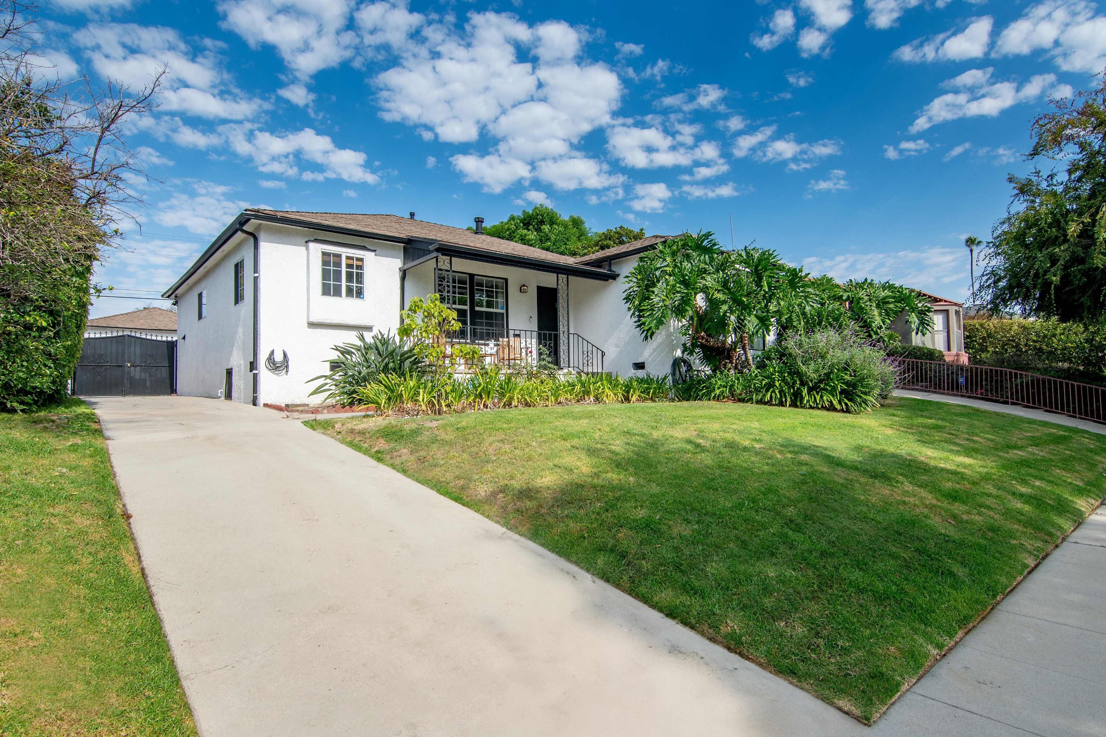 A single-story house with a manicured lawn, surrounded by greenery, sits under a blue sky filled with scattered clouds.