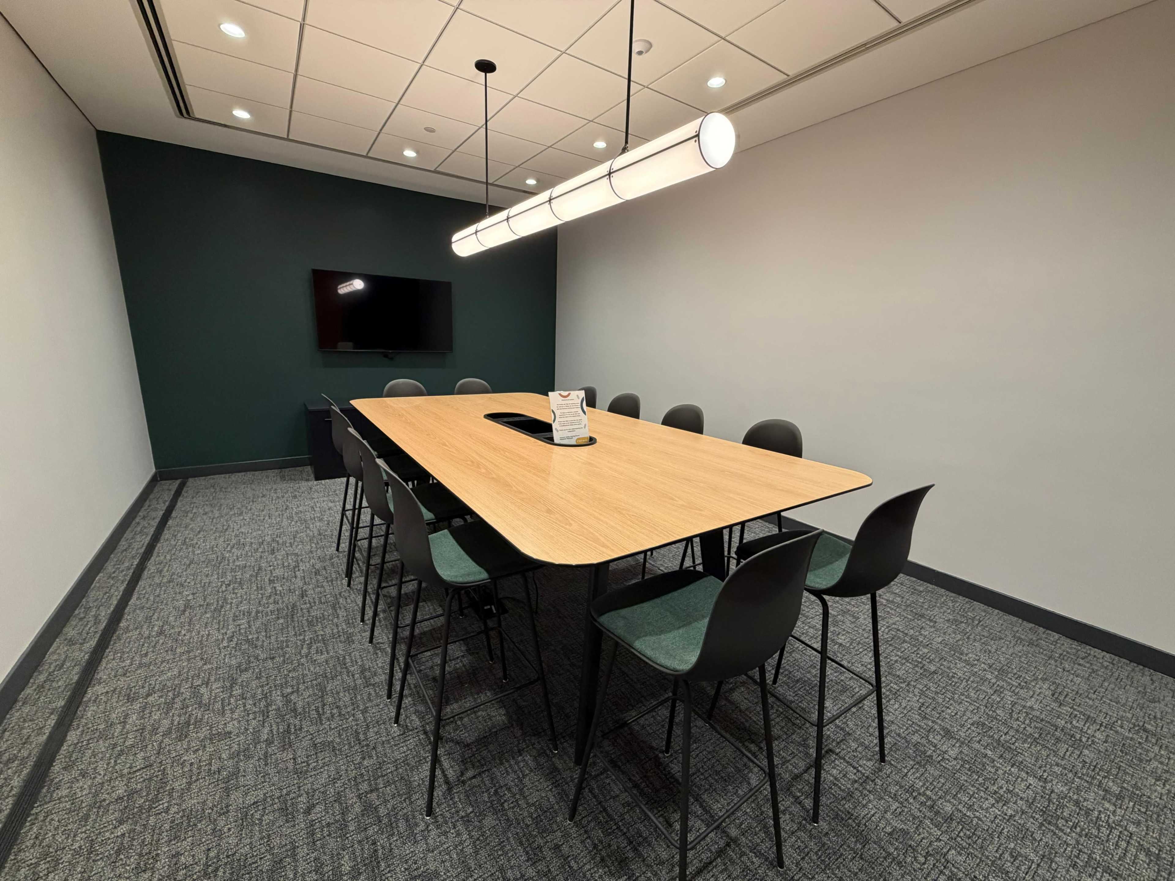 The image shows a modern conference room with a large wooden table surrounded by black chairs, a television mounted on the wall, and a light fixture overhead.