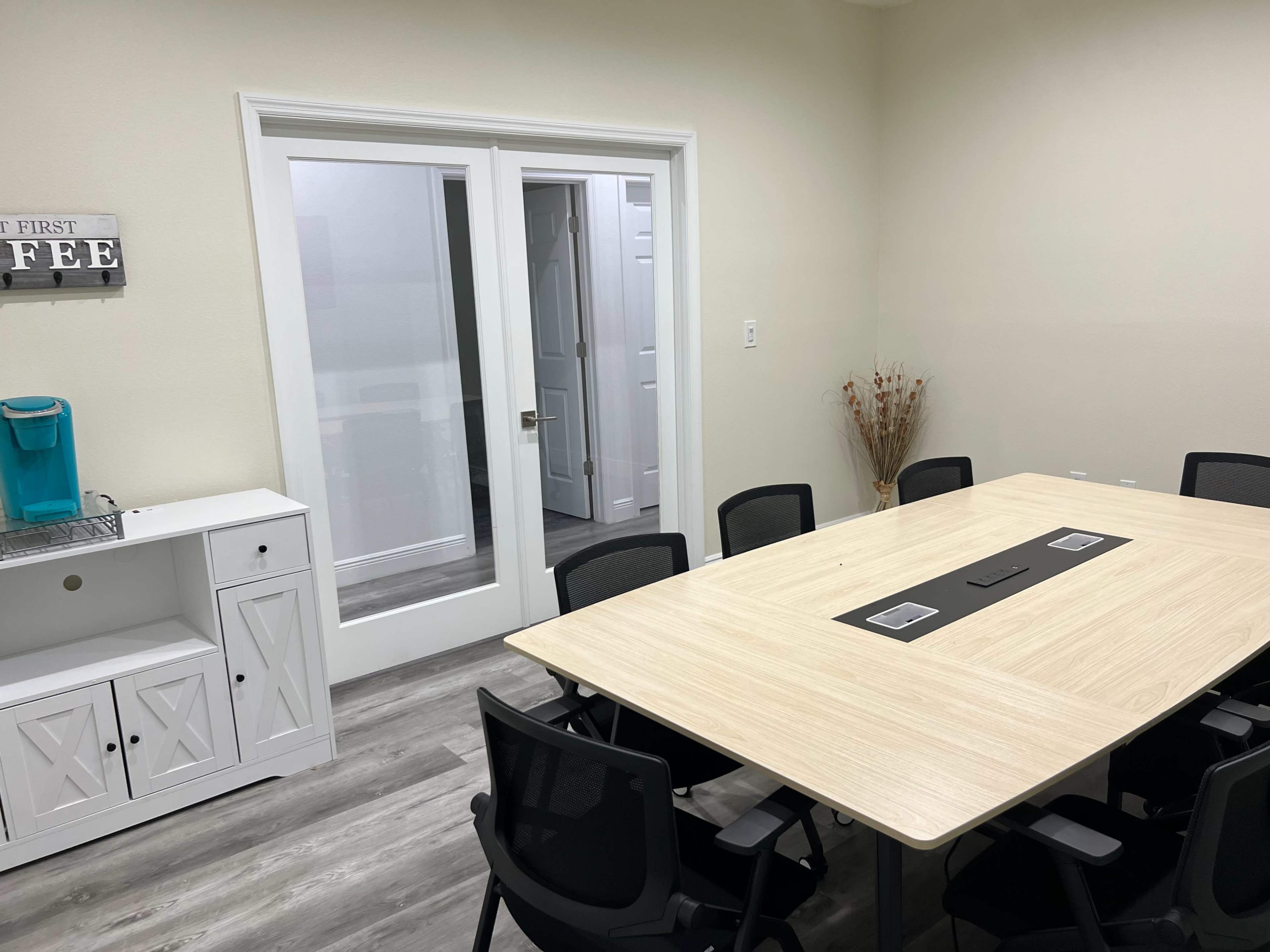 The image shows a modern conference room with a large rectangular table surrounded by chairs, a white cabinet, and a glass door leading to another room.