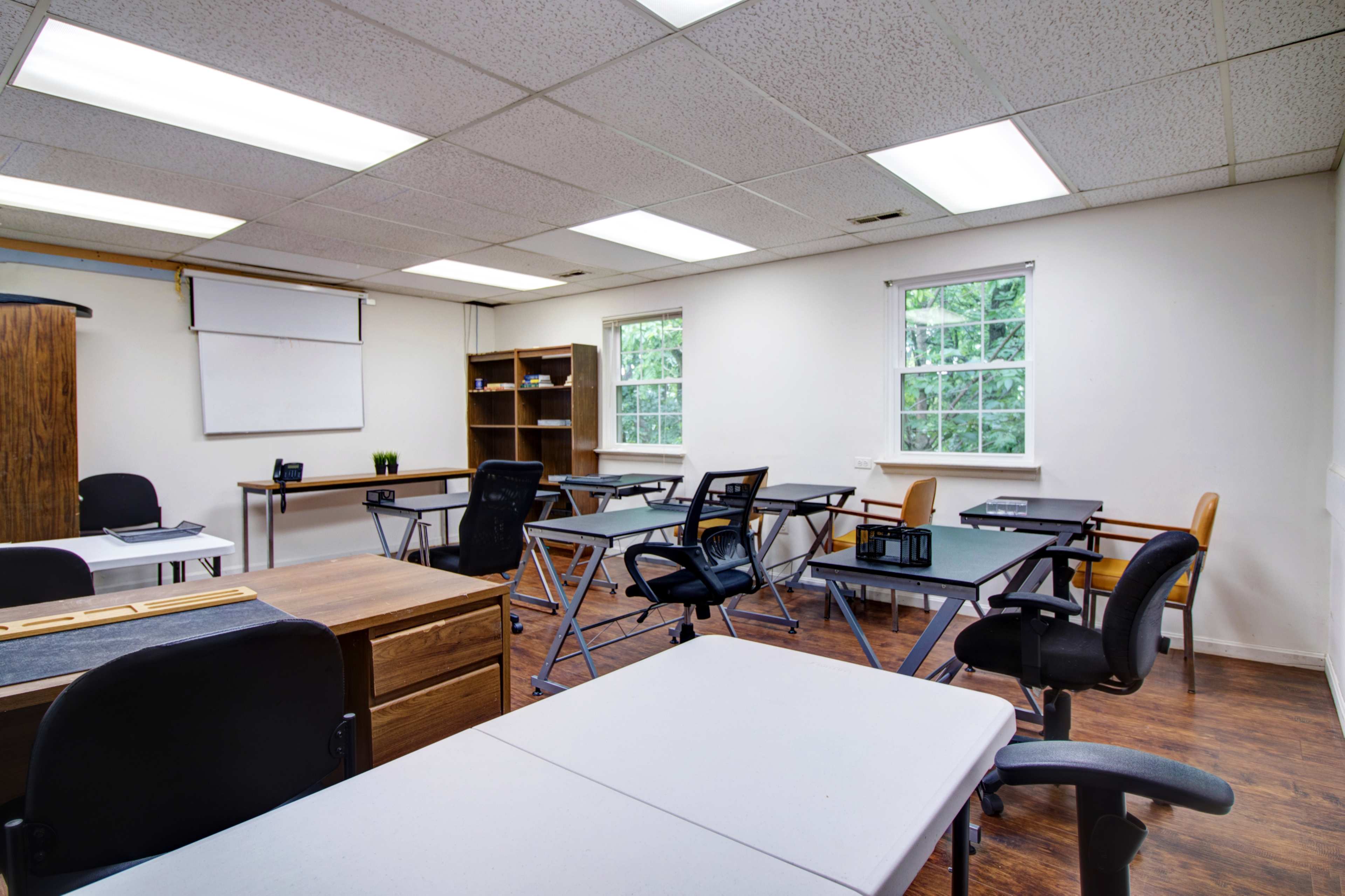 A well-lit classroom with multiple desks, chairs, a whiteboard, and shelves against the walls.