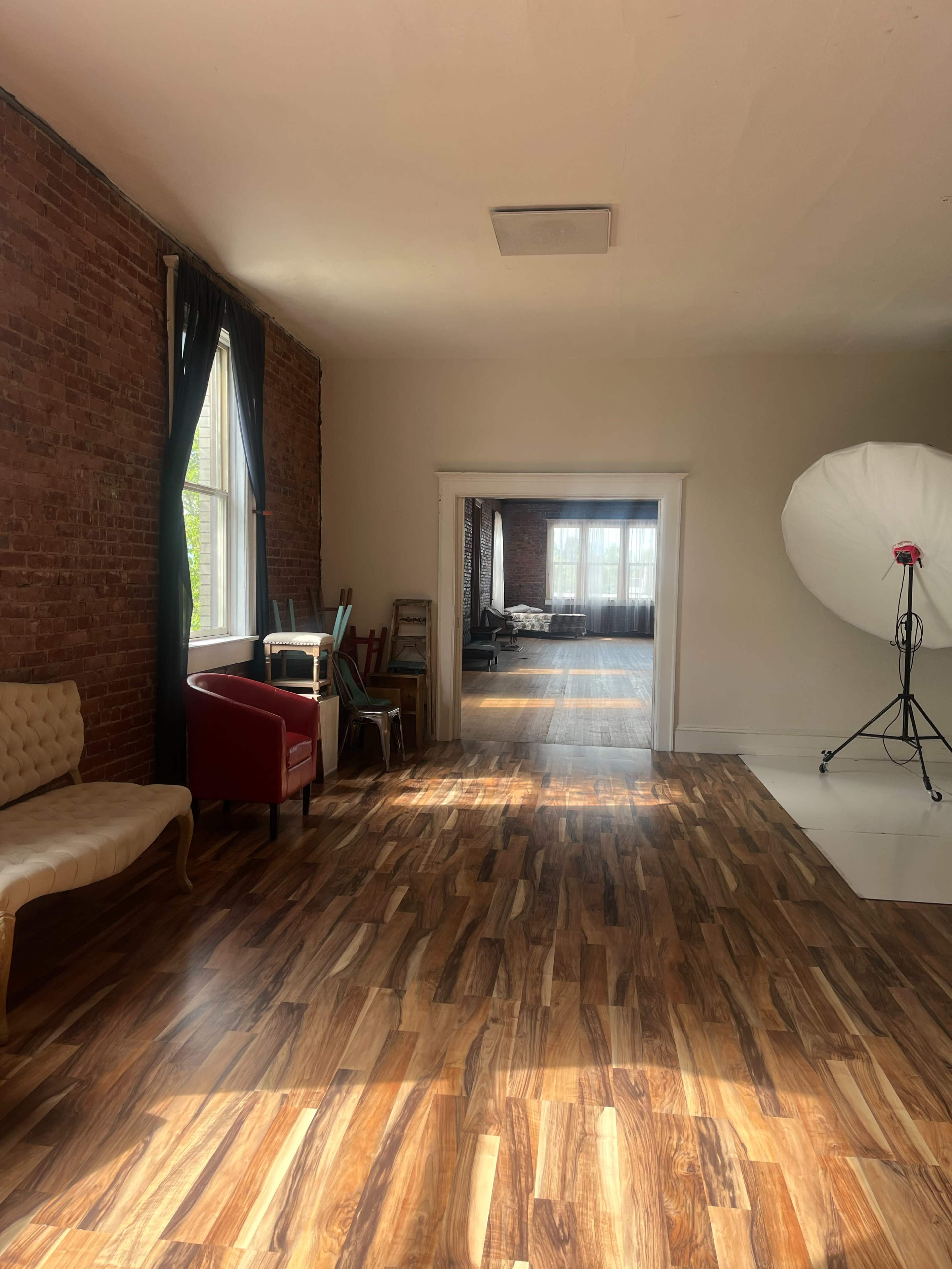 A spacious room with brick walls, hardwood flooring, and a large window, leading to another room filled with natural light and photography equipment.