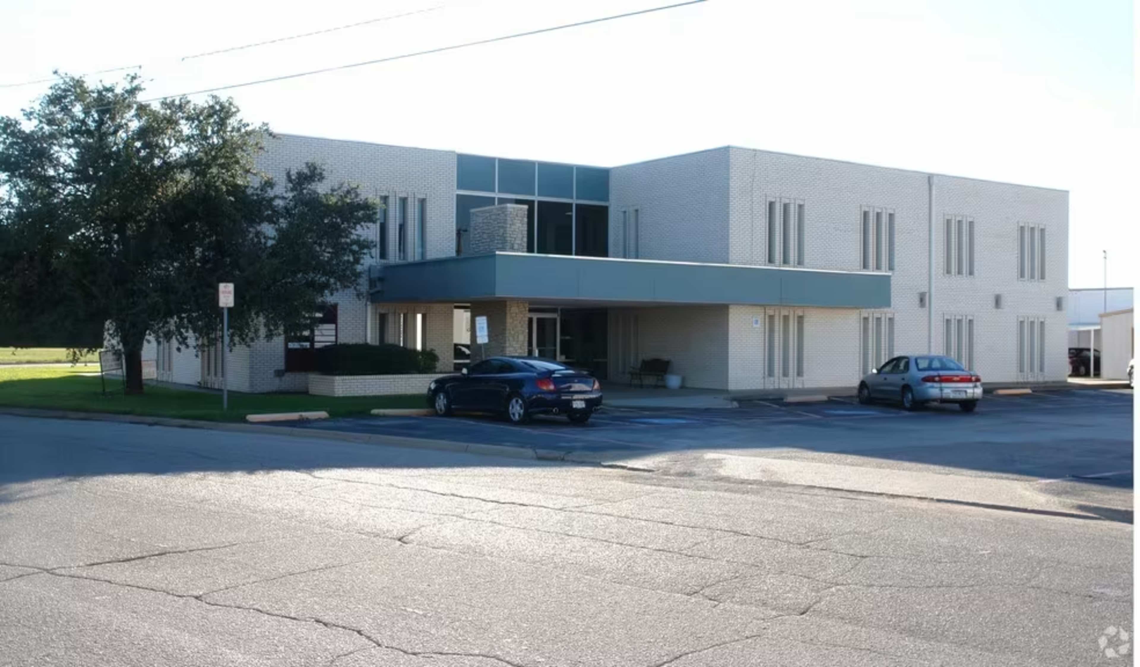 A two-story commercial building with a light-colored brick facade, featuring several windows and two parked cars in the foreground.