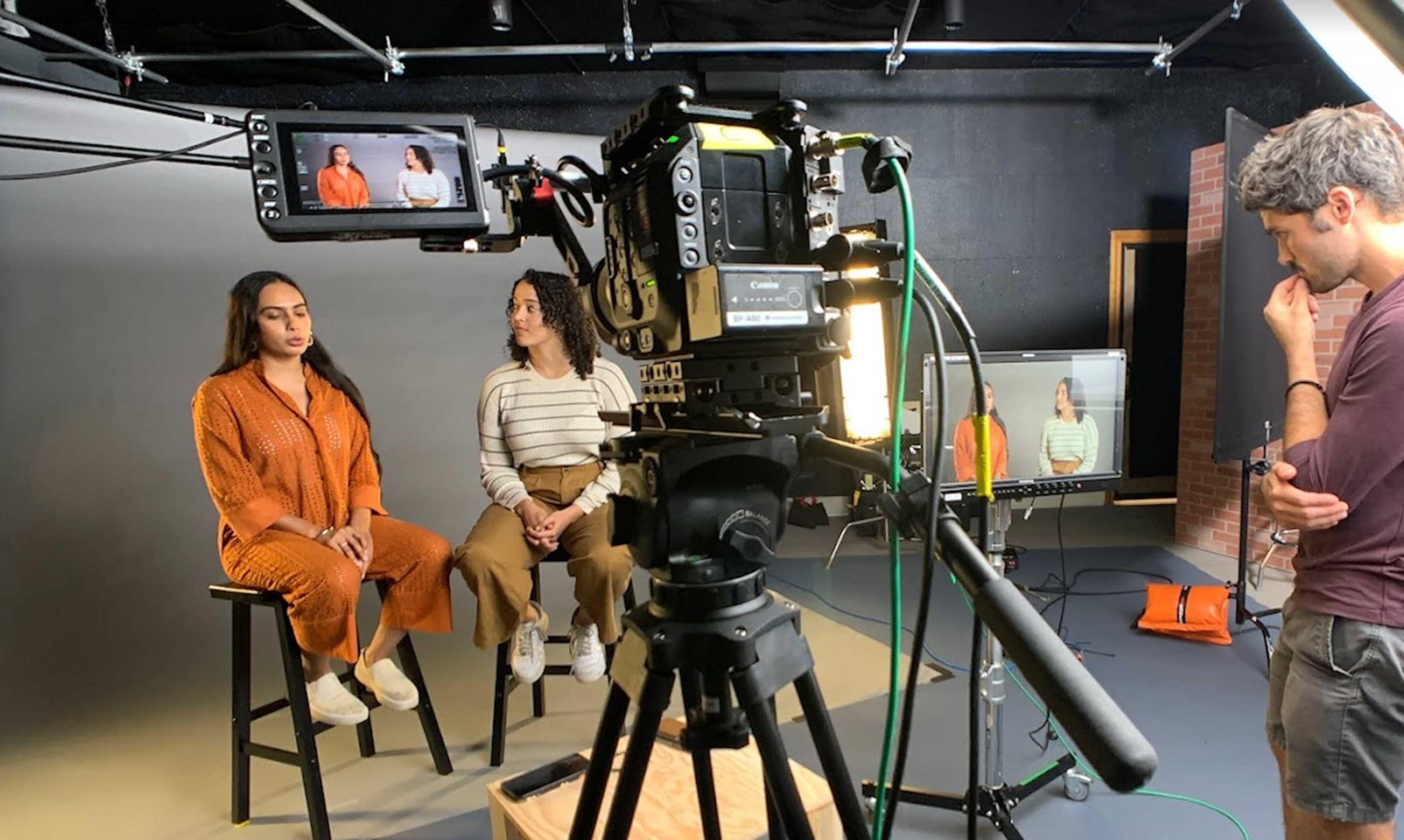 Two women are seated on stools in a studio setting while a man operates a camera on a tripod.