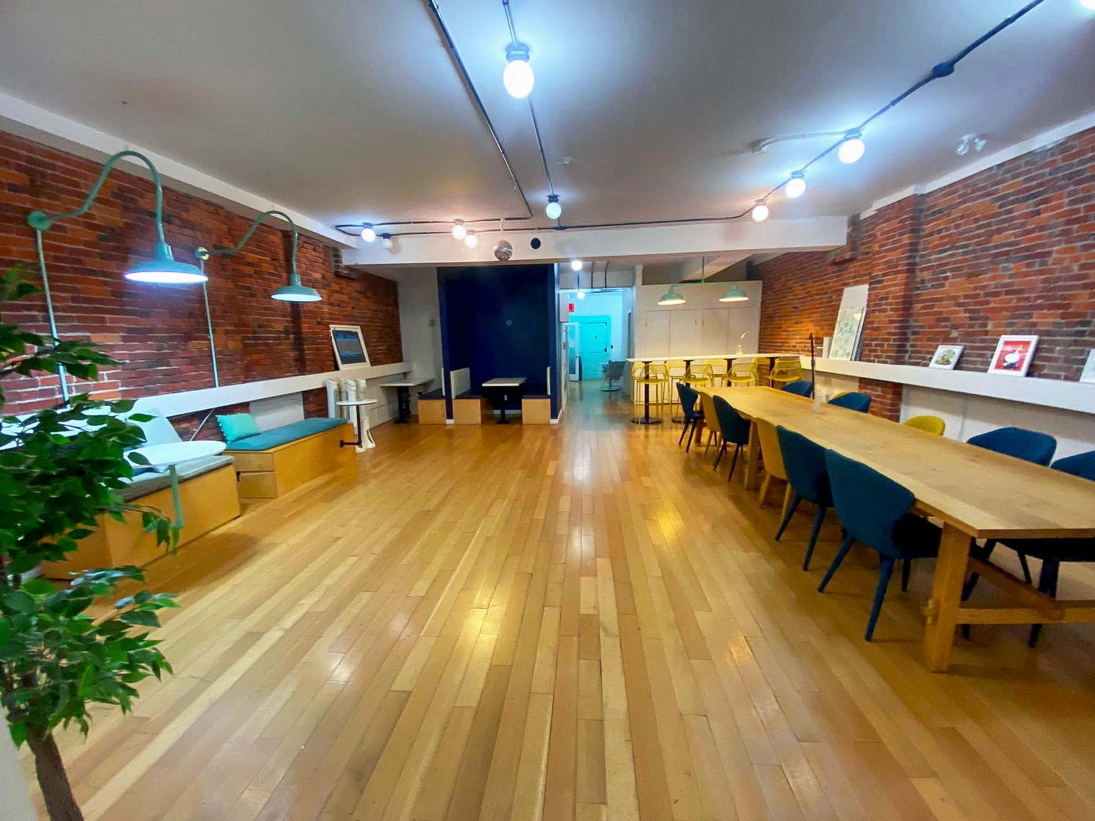 A spacious, well-lit room with wooden floors, exposed brick walls, a long dining table, seating areas, and modern light fixtures.