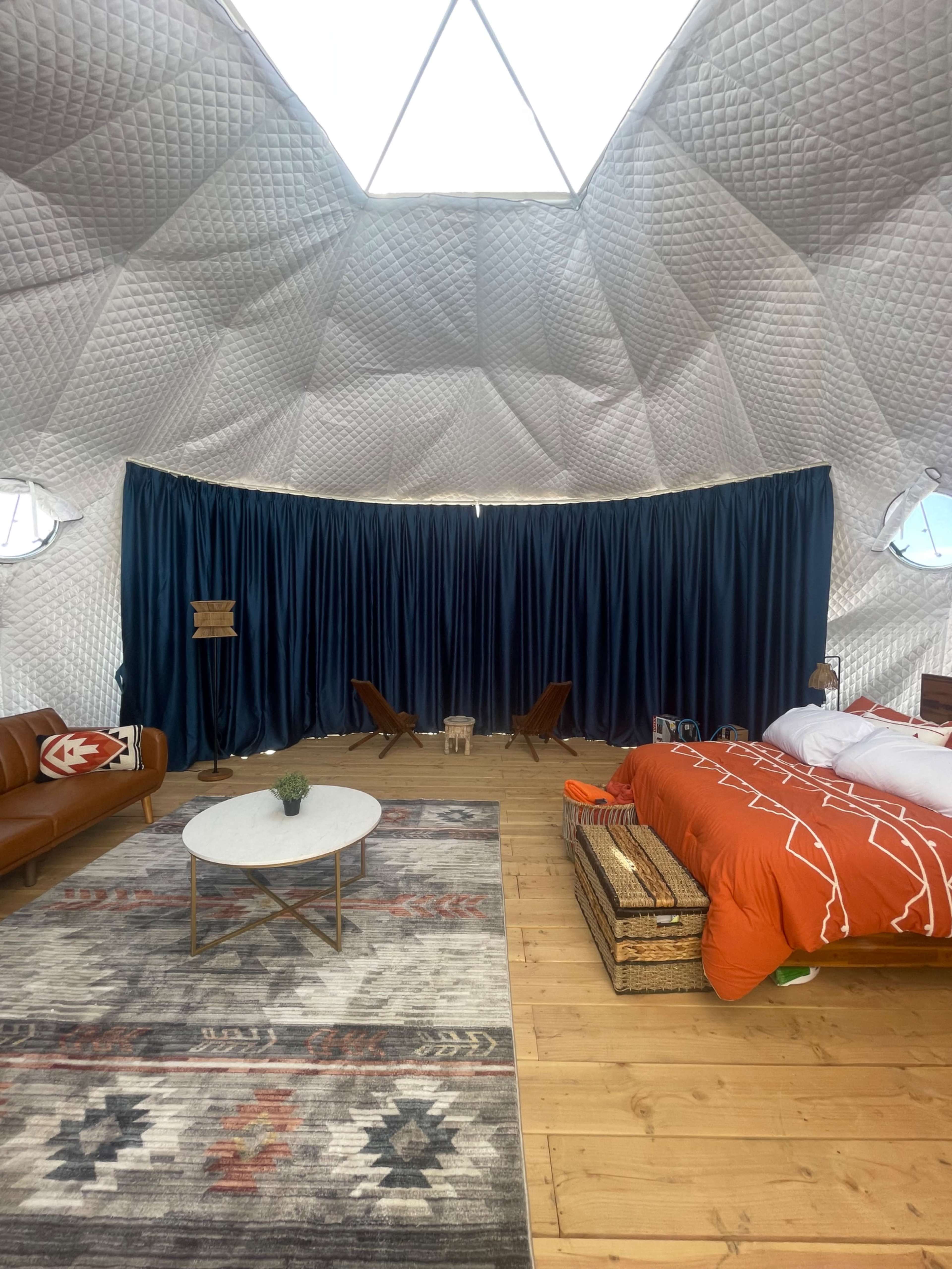 A spacious, modern bedroom with a domed ceiling, featuring a large bed, a circular coffee table, and a seating area with two armchairs, all surrounded by soft lighting and curtains.