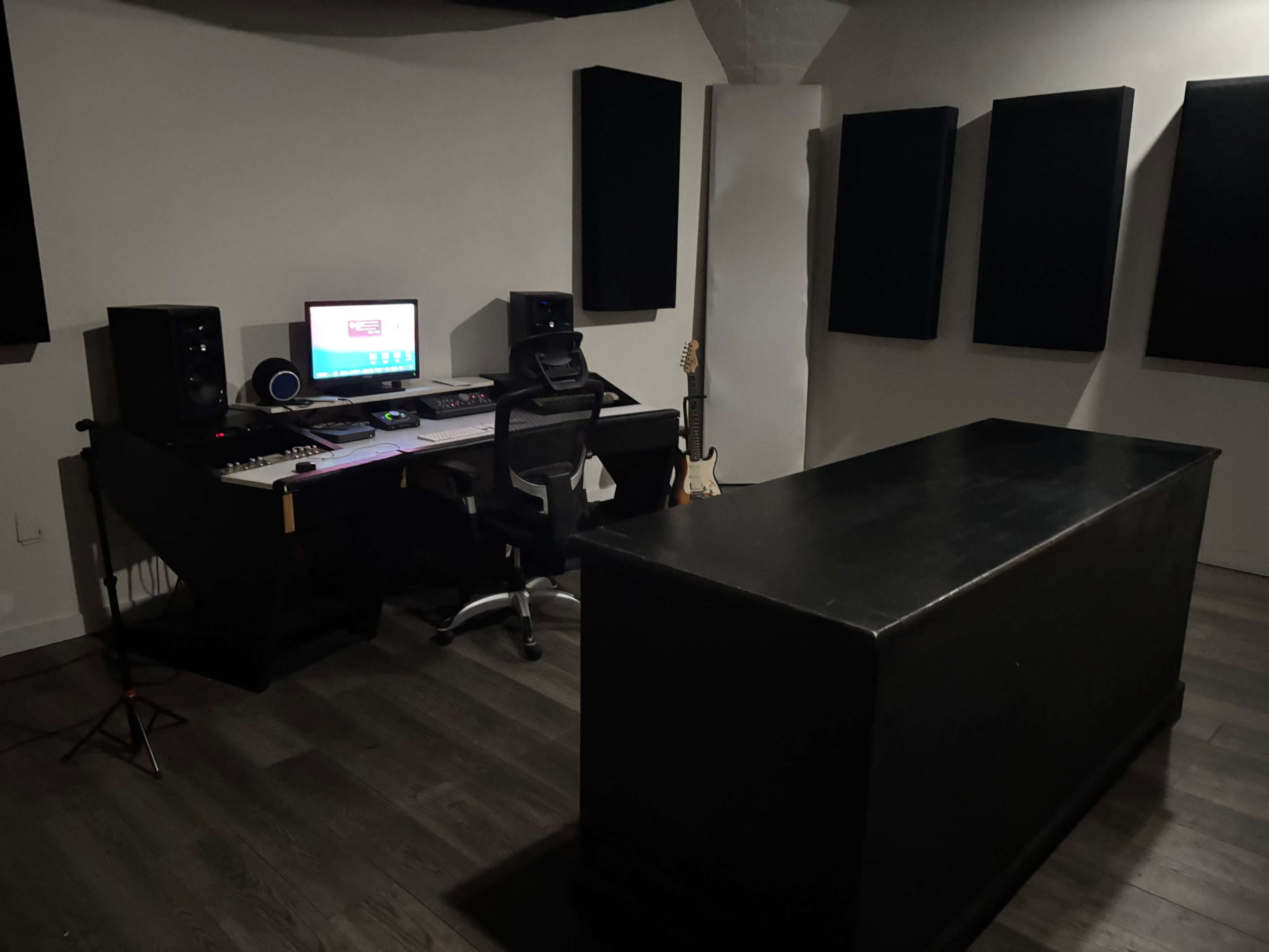 The image shows a music studio with a desk featuring a computer, speakers, and an office chair, alongside a black console and an electric guitar in the corner.
