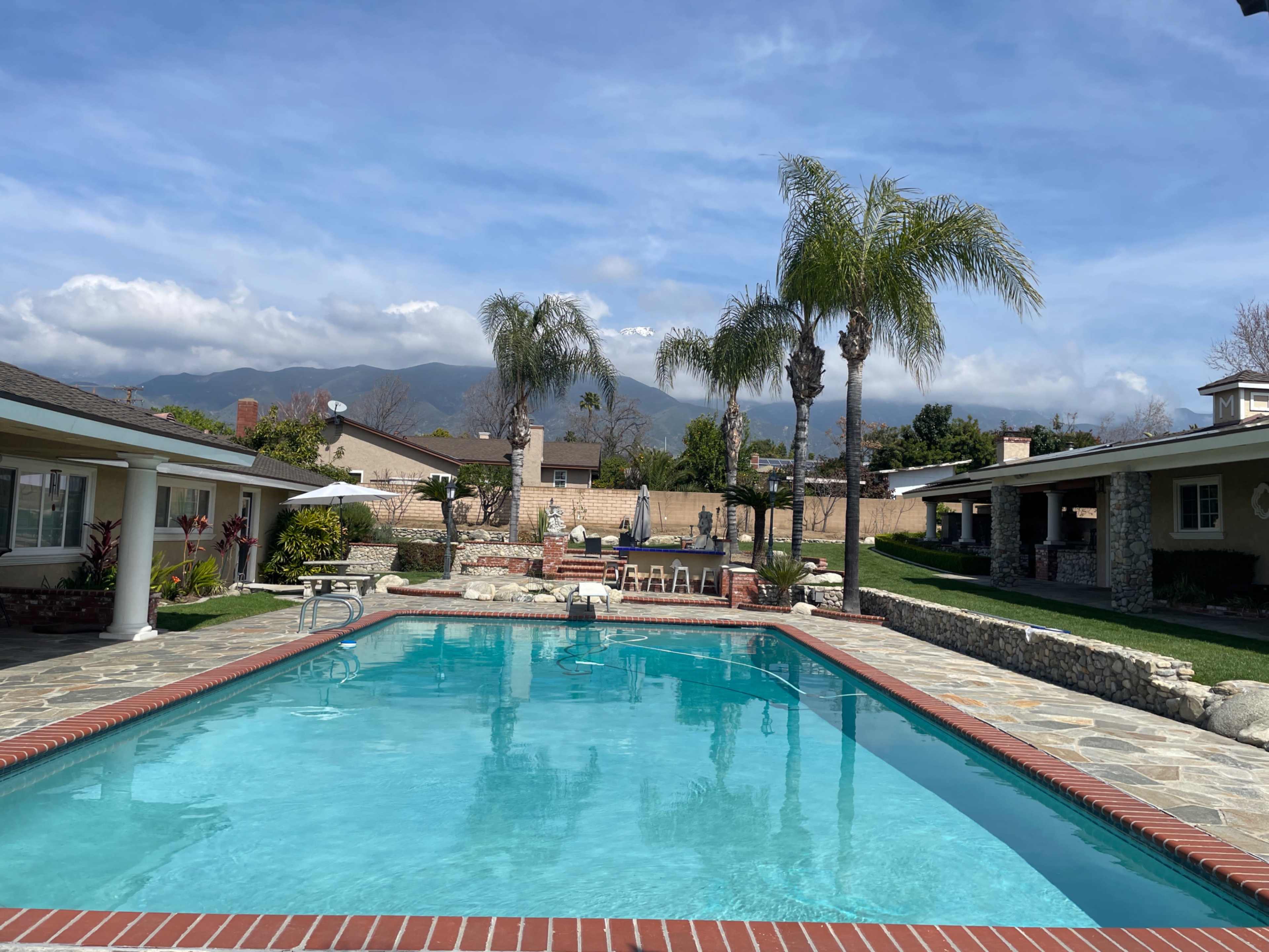 A clear swimming pool is surrounded by palm trees and houses, with mountains visible in the background under a partly cloudy sky.