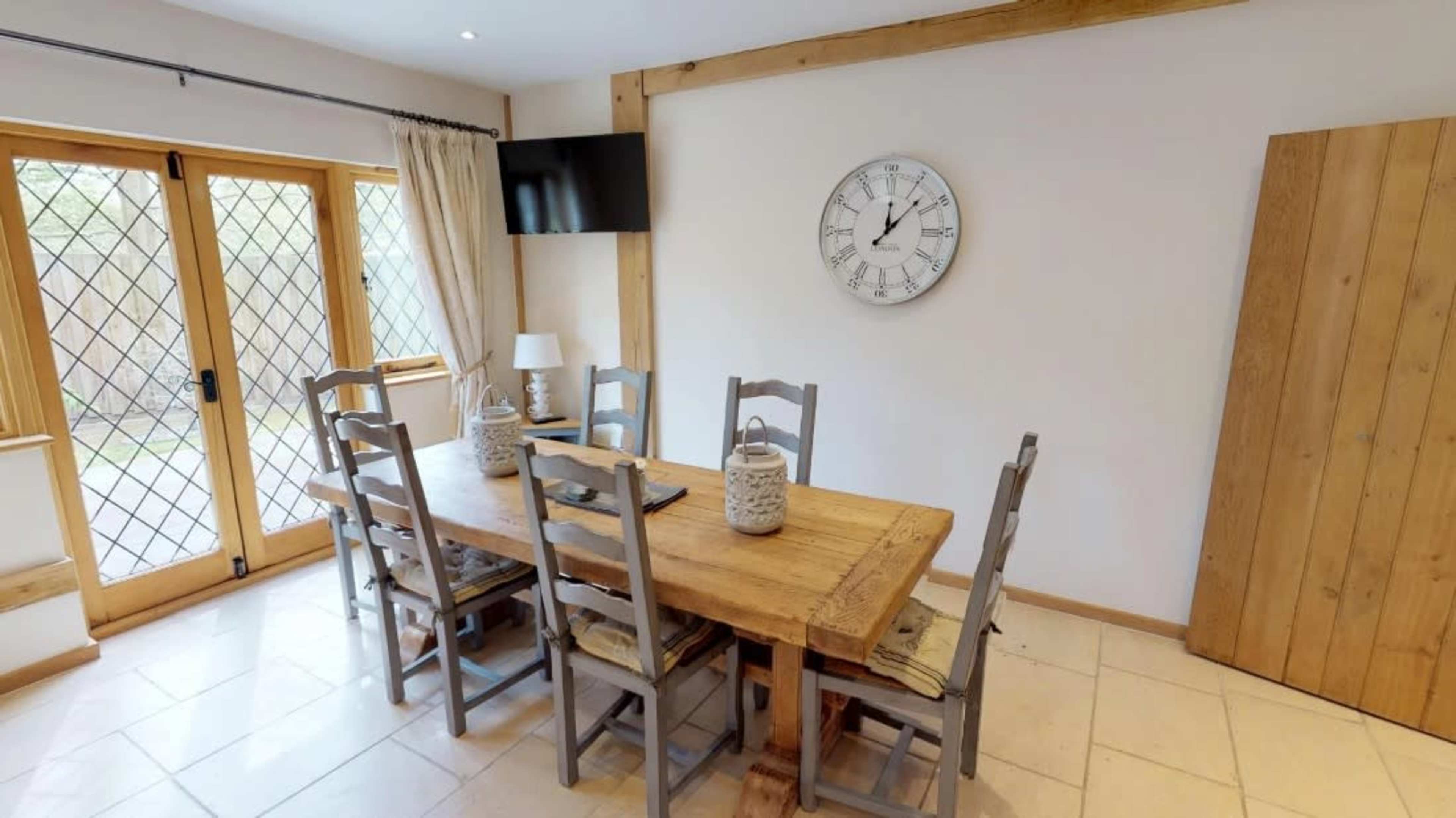 A wooden dining table surrounded by six chairs is positioned in a room with large windows and a wall clock.