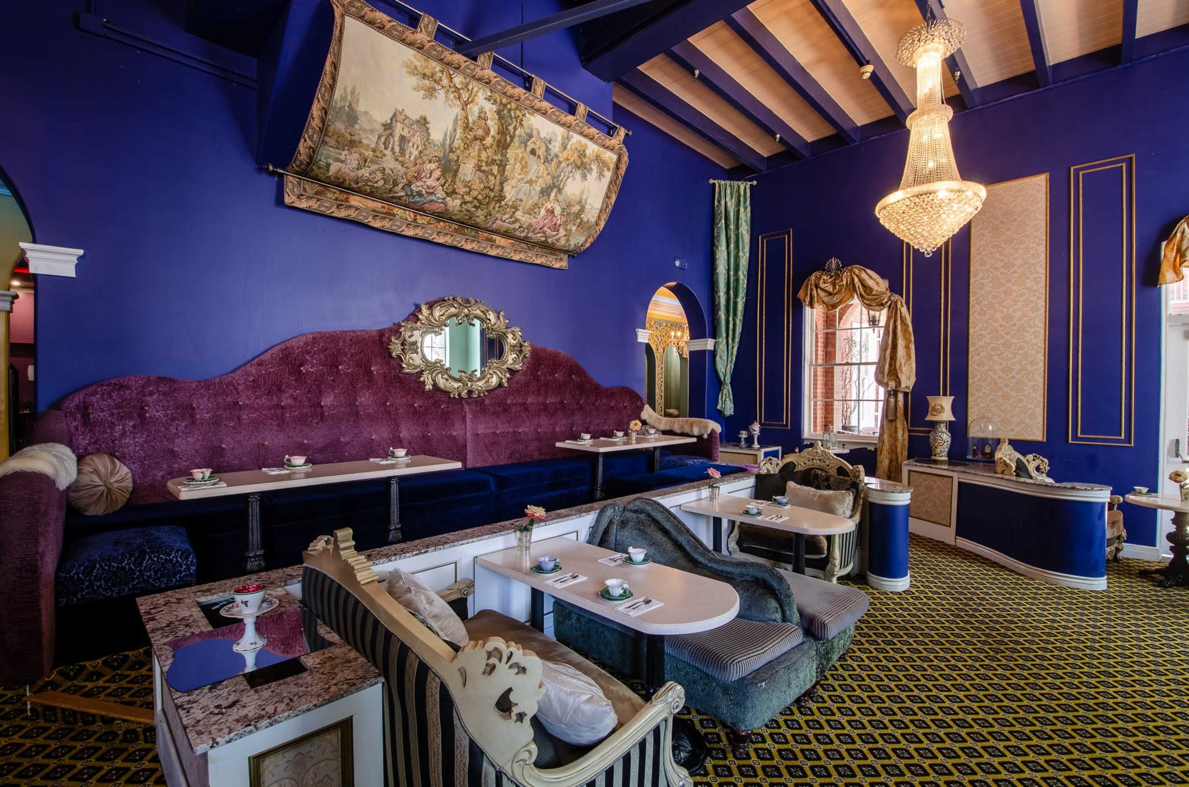 The image shows a vibrant lounge with purple walls, ornate furniture, and a crystal chandelier, featuring several small tables arranged around plush seating areas.
