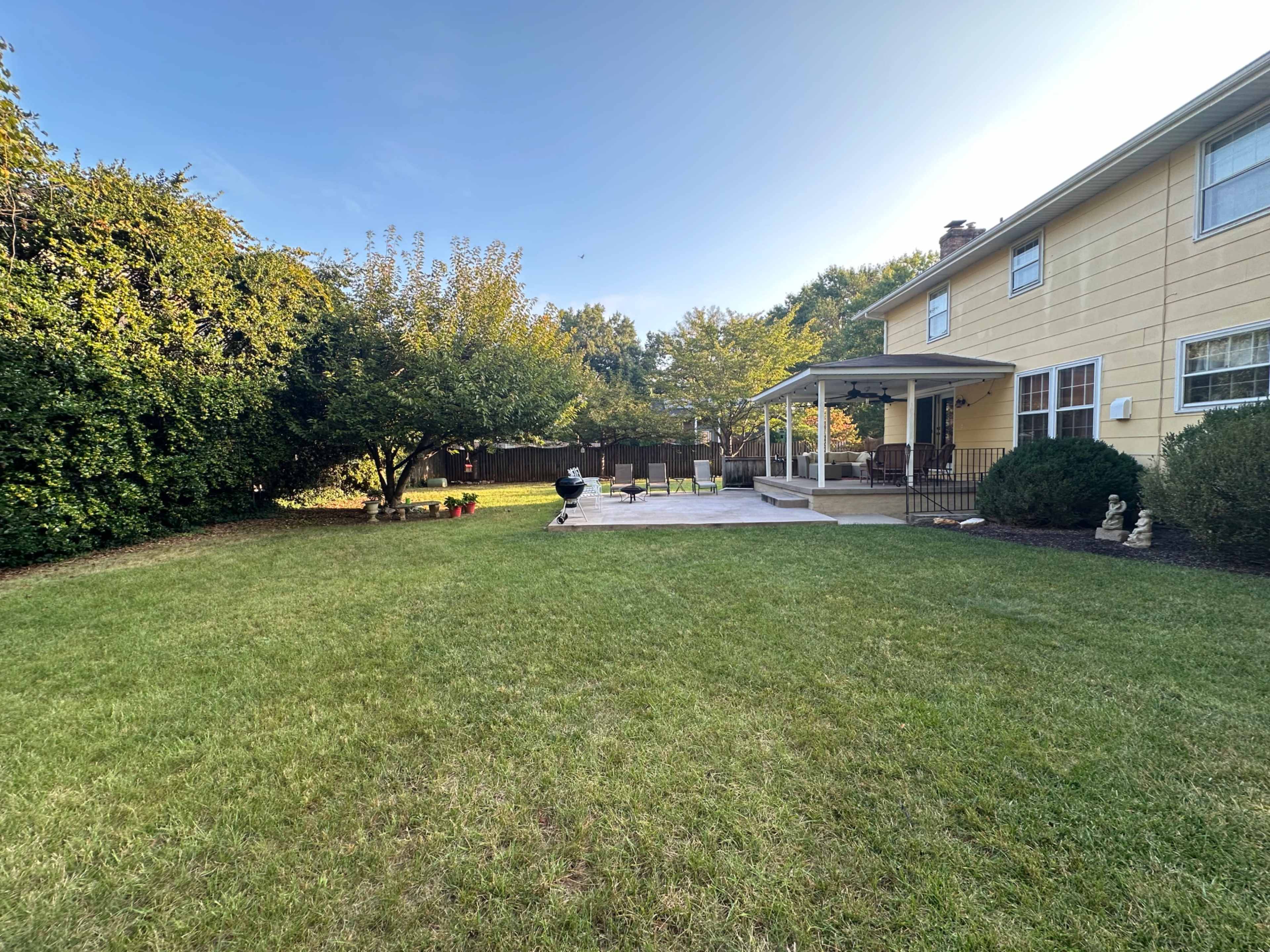 A grassy yard is seen with a patio, a house in the background, and a person standing near a grill.
