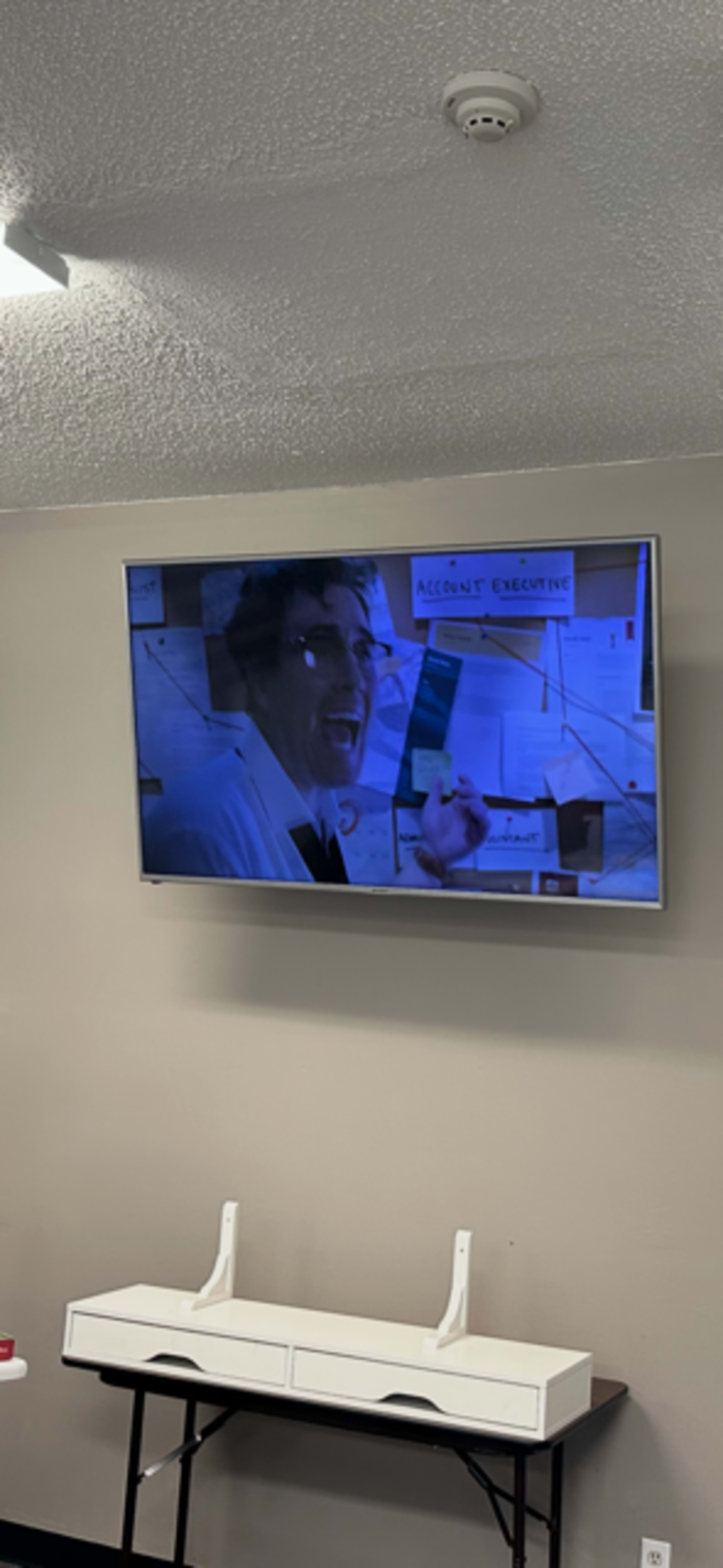 A television mounted on the wall displays a scene featuring a man excitedly gesturing while seated at a desk in an office setting.