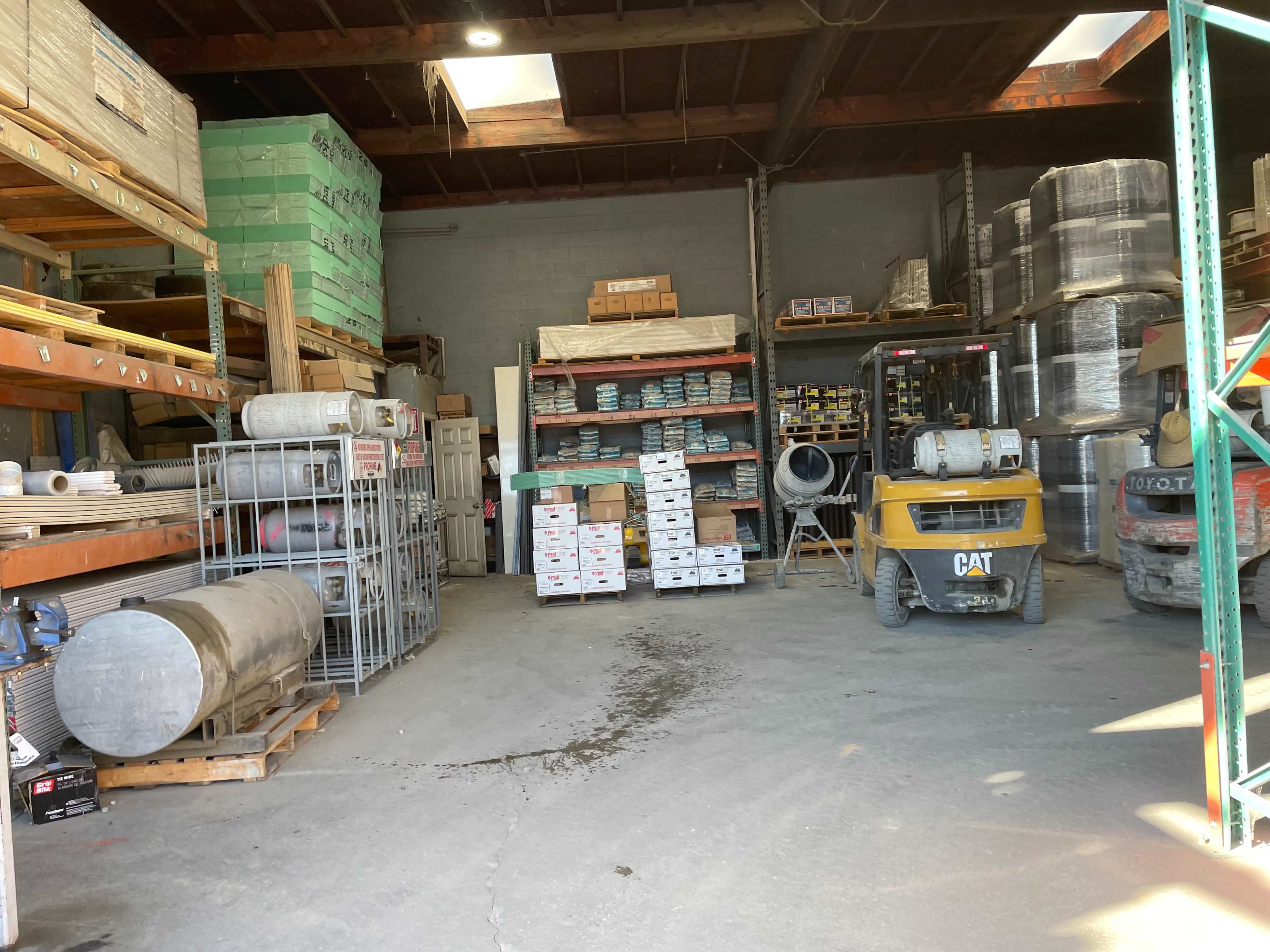 The image shows a warehouse interior with shelves stocked with various construction materials, equipment, and a forklift.