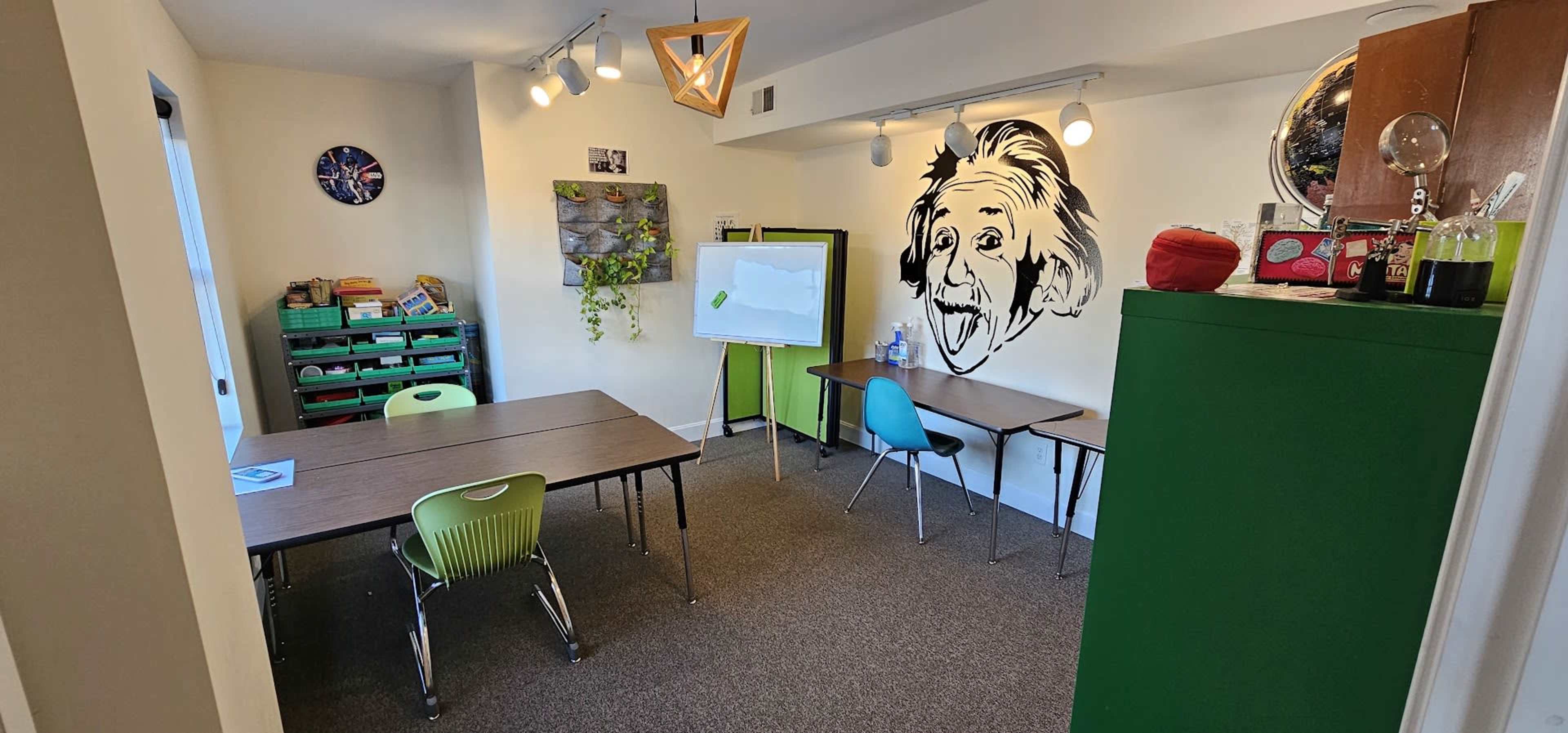 A classroom features several tables and chairs, a whiteboard, and a wall mural of Albert Einstein.