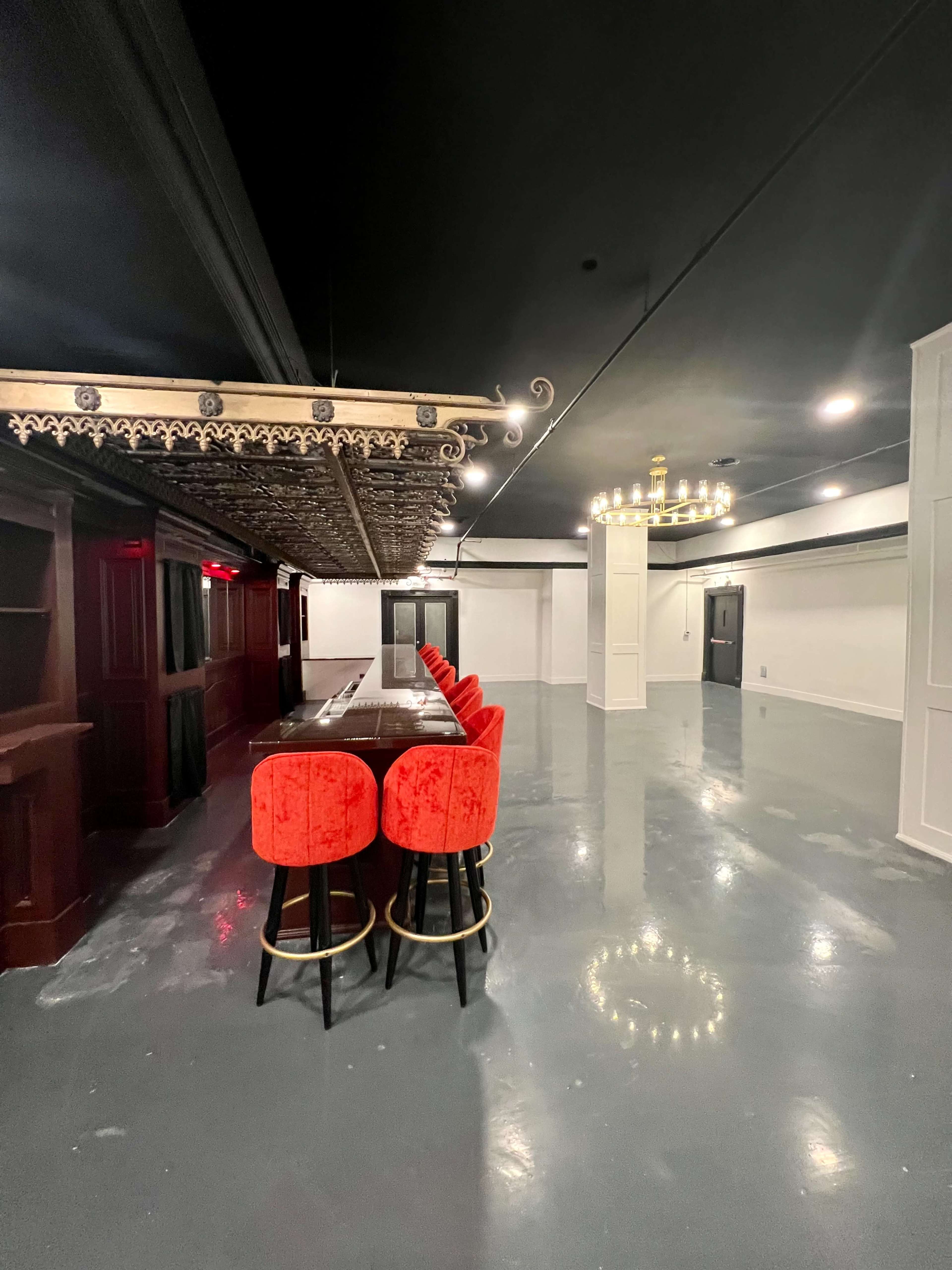 A spacious room with a long dark table, red upholstered stools, ornate ceiling details, and a chandelier.