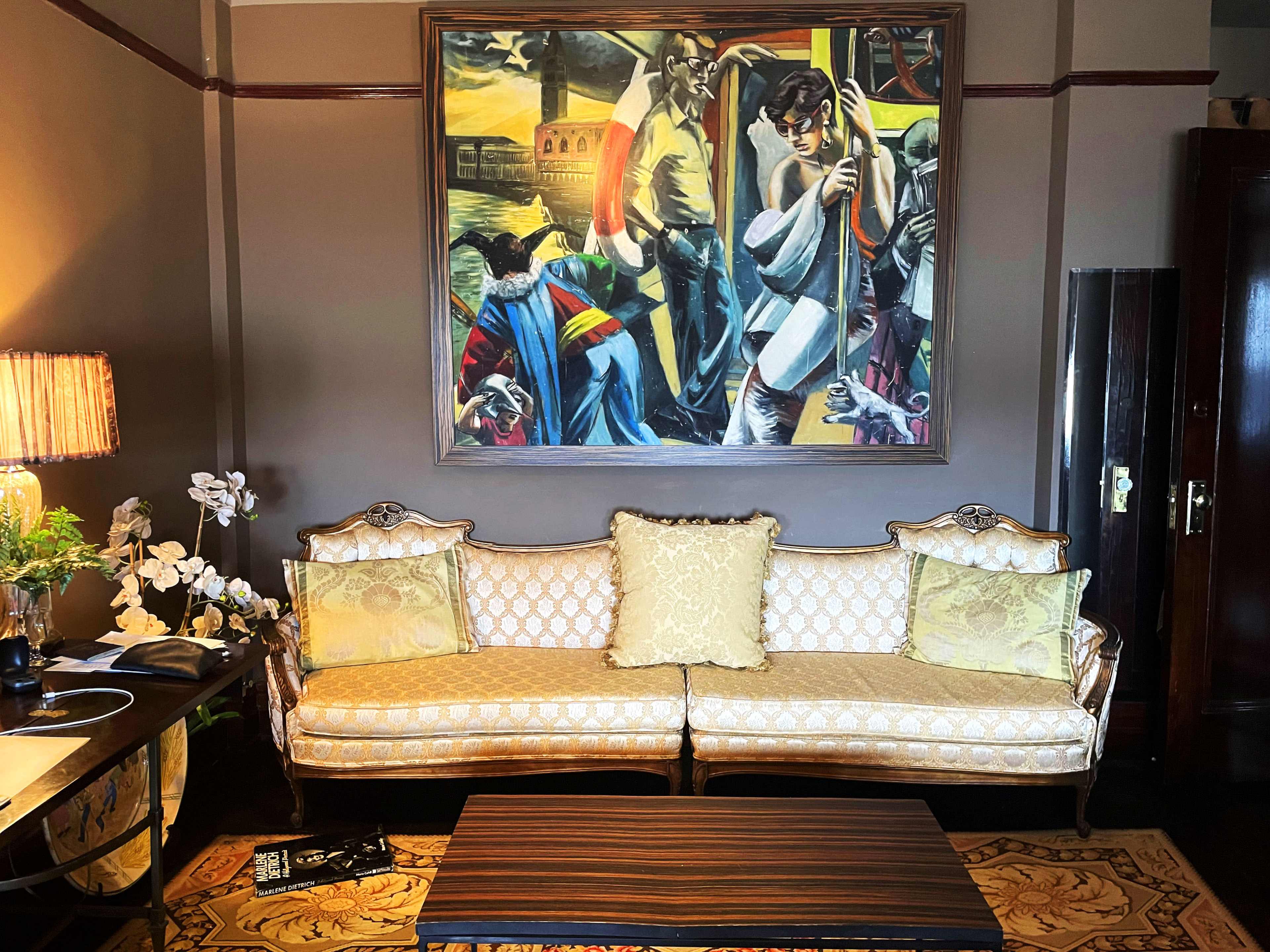 A vintage living room with a patterned sofa and a large painting on the wall depicting a scene with figures, alongside a coffee table and decorative elements.