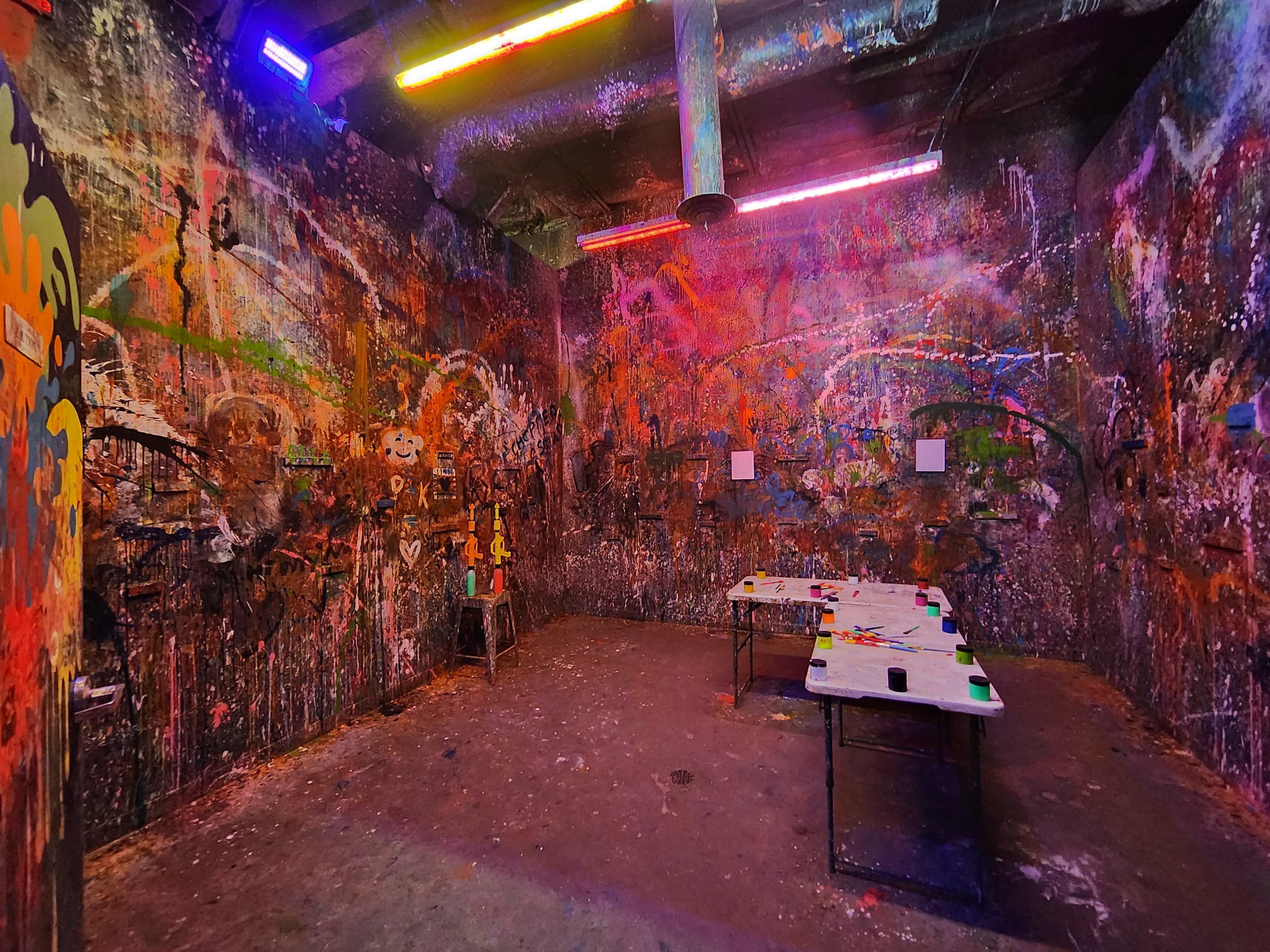 The image shows a dimly lit room with vibrant, multicolored paint splatters covering the walls and a couple of tables in the center.