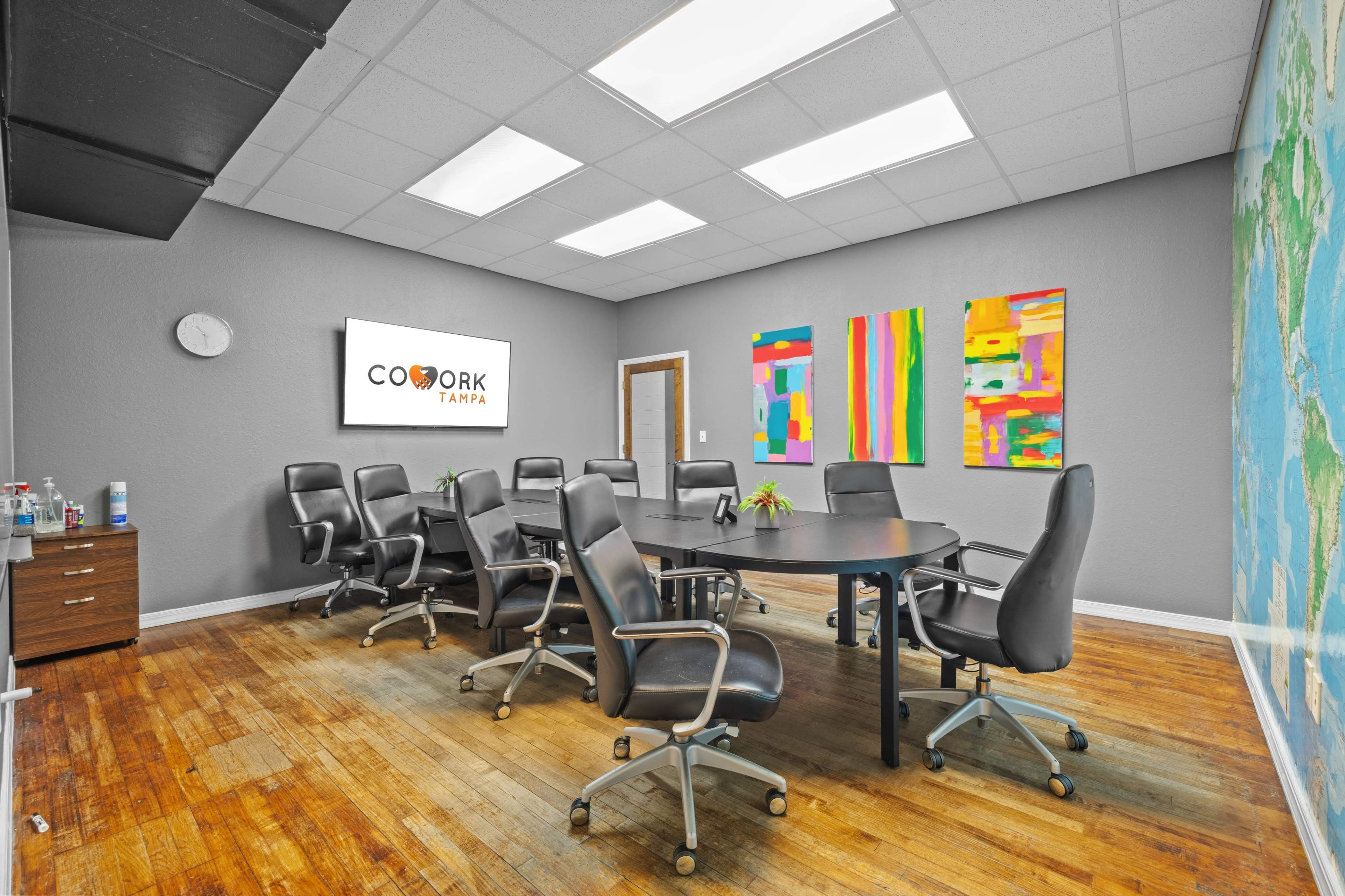 The image shows a conference room with a round table surrounded by black office chairs, colorful artwork on the walls, and a sign for "COWORK TAMPA."