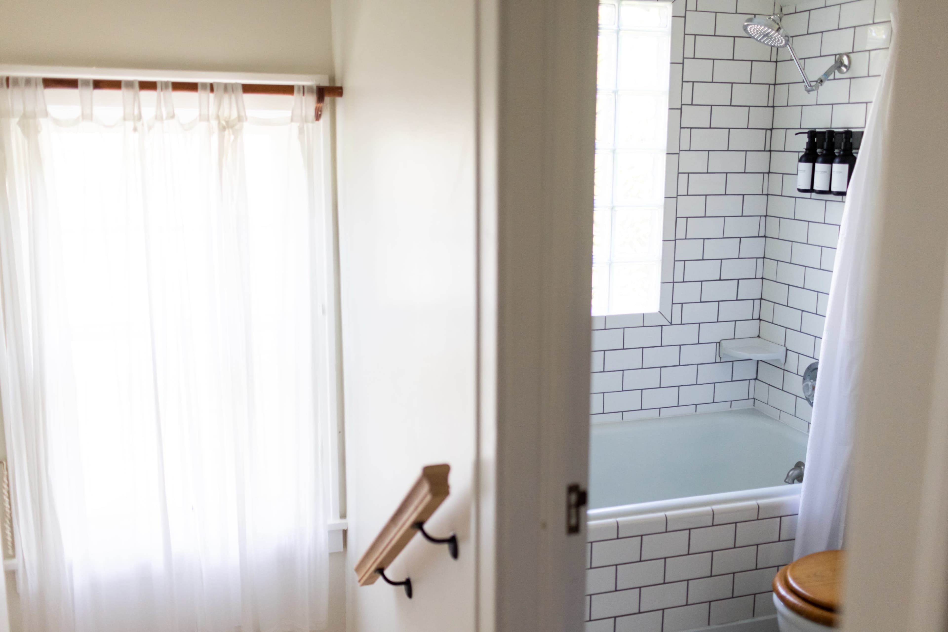 A modern bathroom features a white tiled shower area with a bathtub and a frosted window, while a sheer curtain partially drapes the entrance.