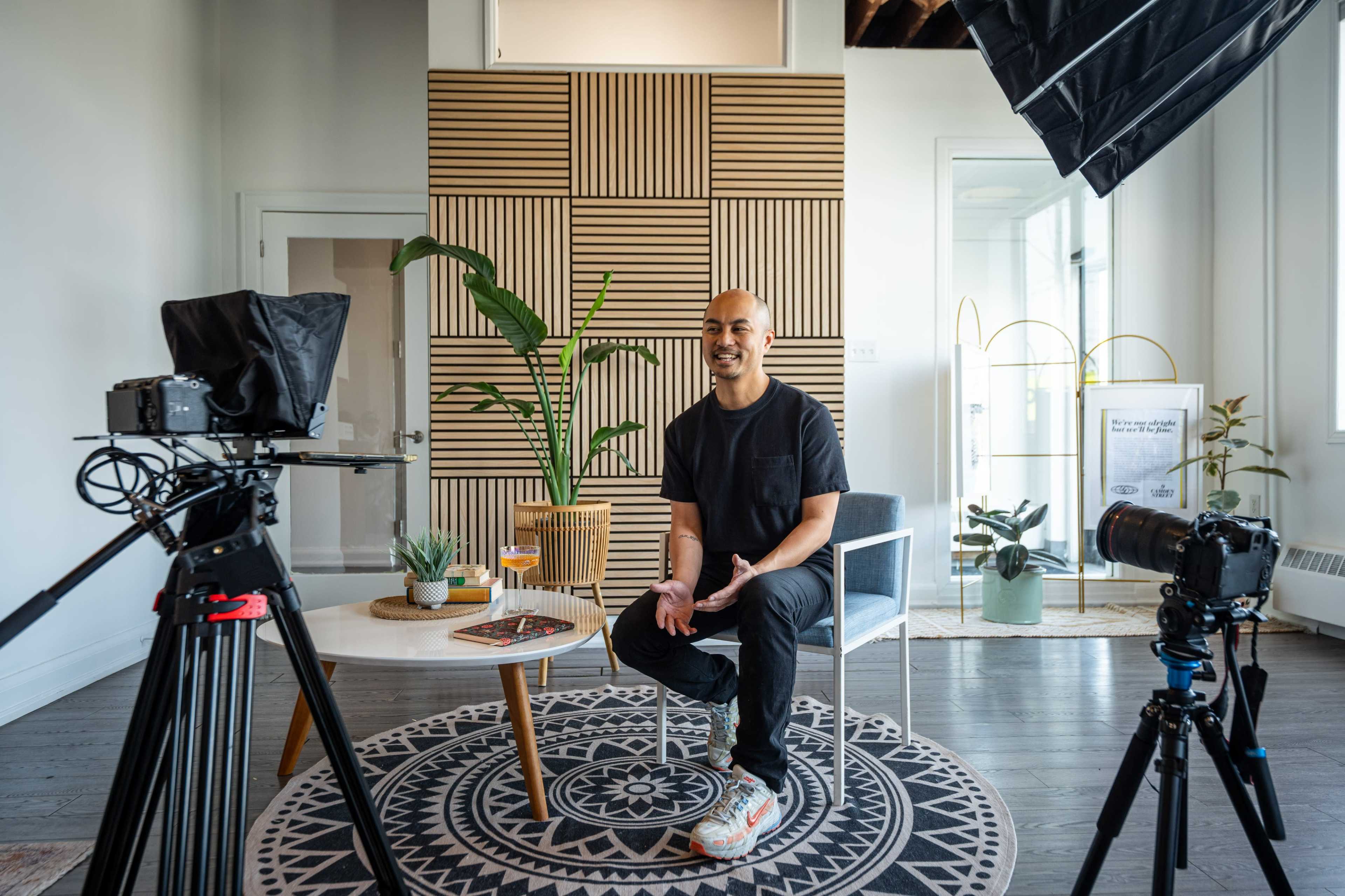 A man sits on a chair in a well-lit room, surrounded by cameras and plants, with a geometric-patterned wall in the background.