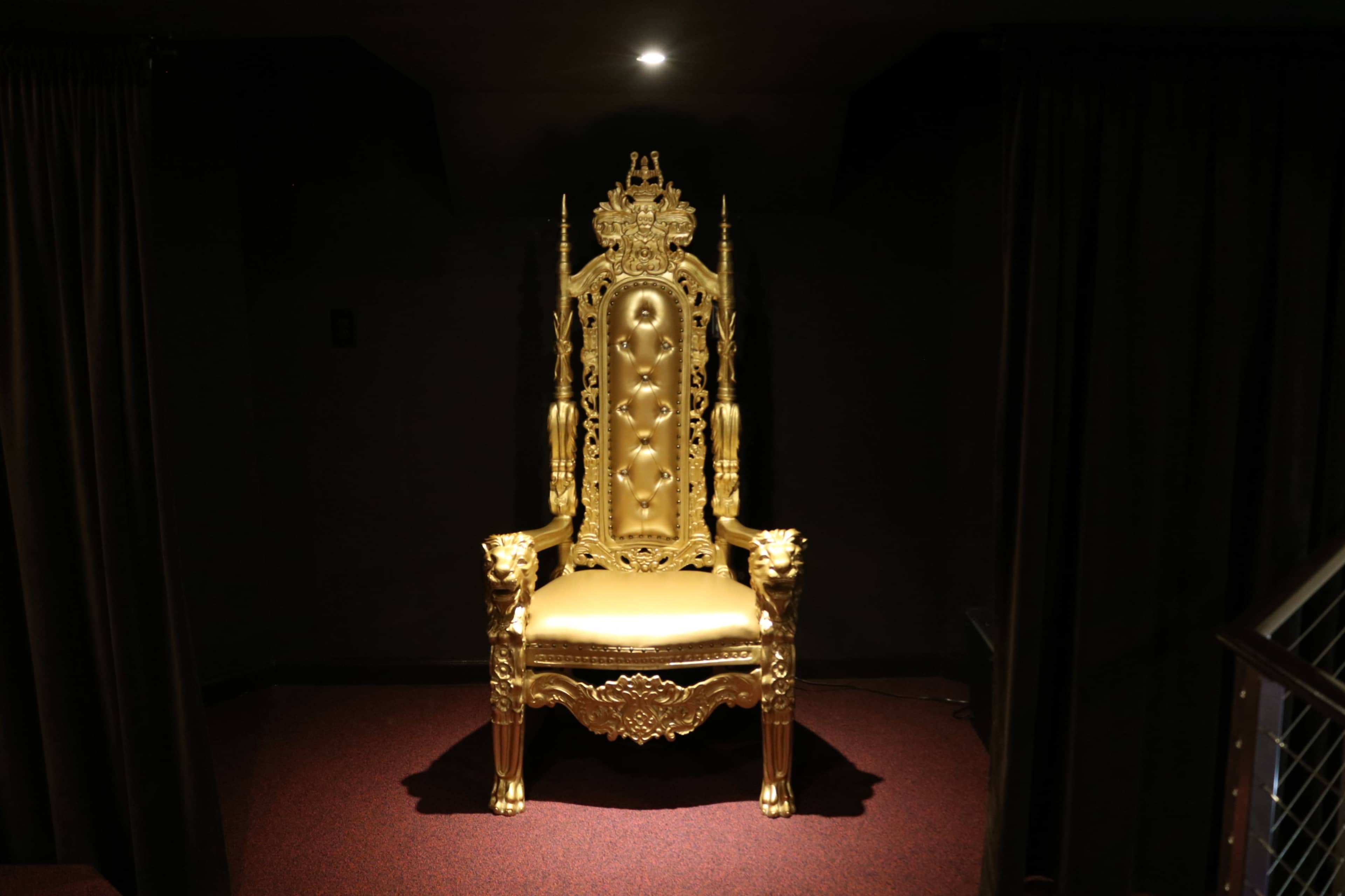 A gold ornate throne with a high back and decorative details is positioned on a red carpeted floor surrounded by dark curtains.
