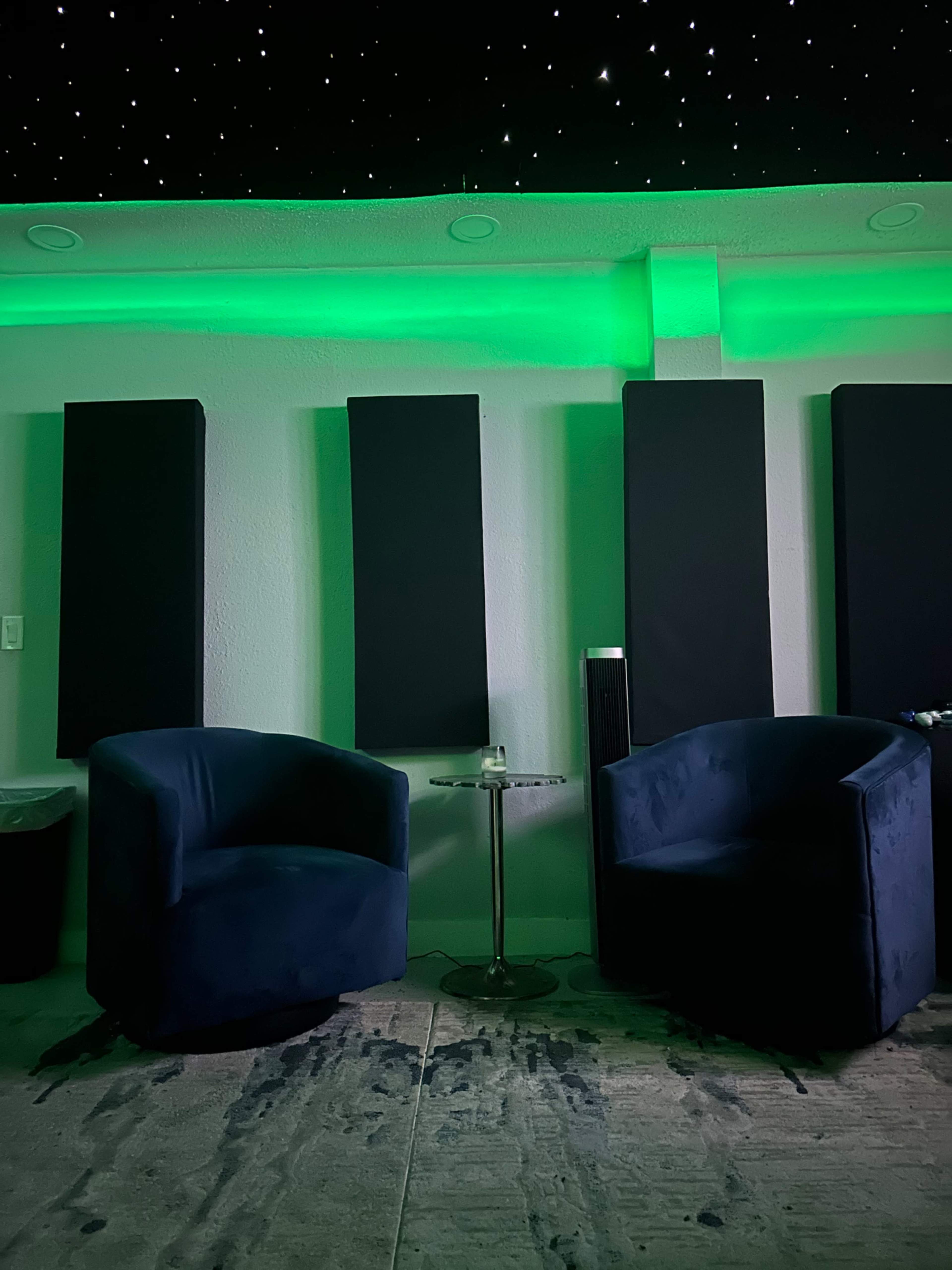 Two blue armchairs positioned around a small glass table under a starry ceiling, with green lighting illuminating the area.