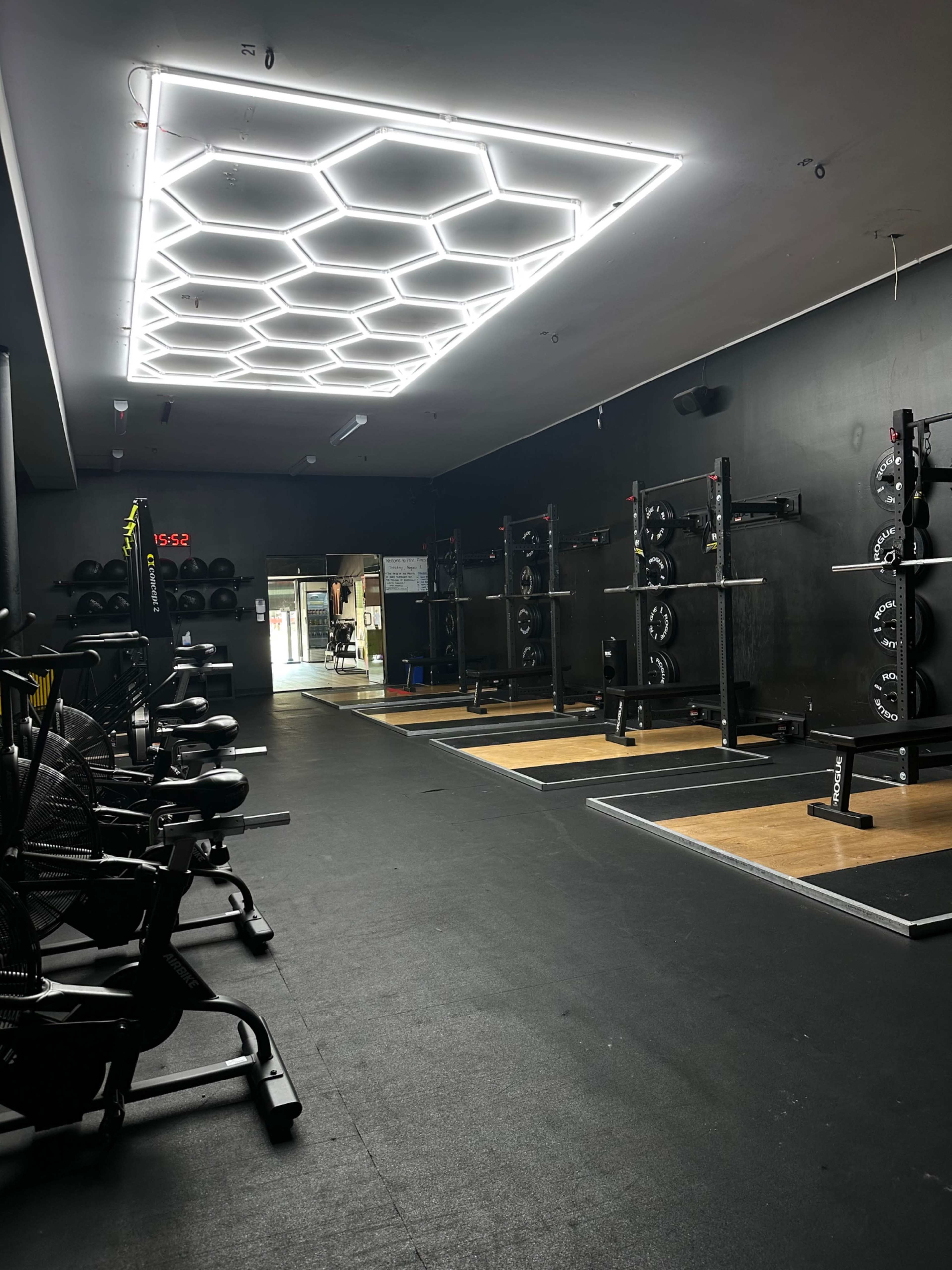 A gym interior with workout stations, including weightlifting racks and exercise bikes, illuminated by a hexagonal LED ceiling light.