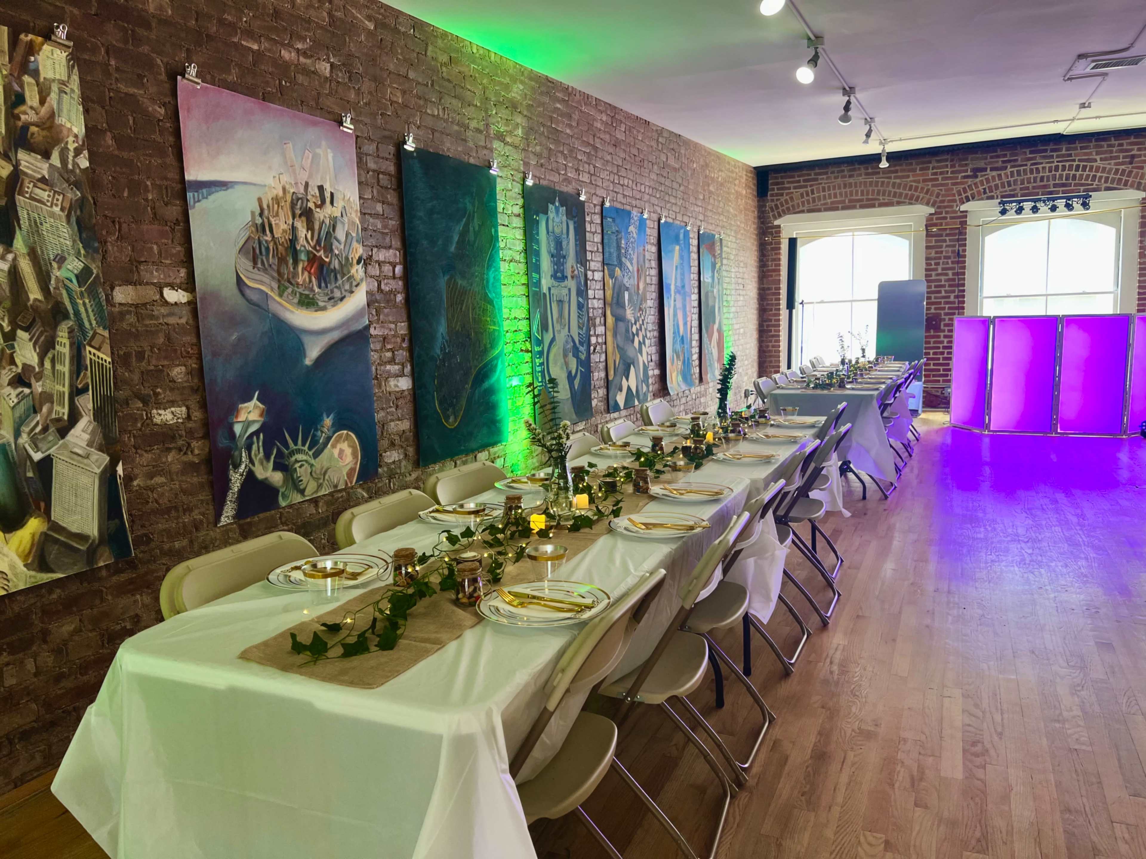 A long dining table is set up in a brick-walled room adorned with artwork, with greenery and candles as centerpieces.