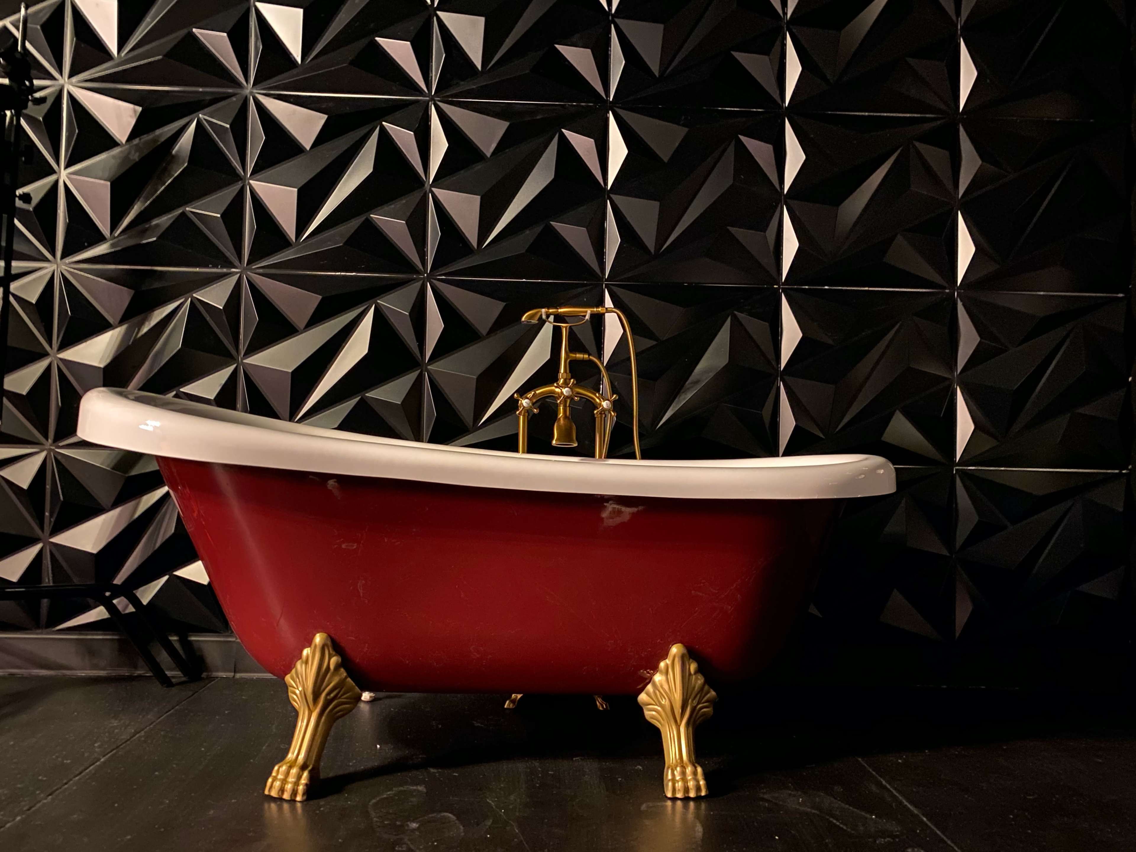 A vintage red bathtub with gold fixtures sits against a black geometric textured wall.