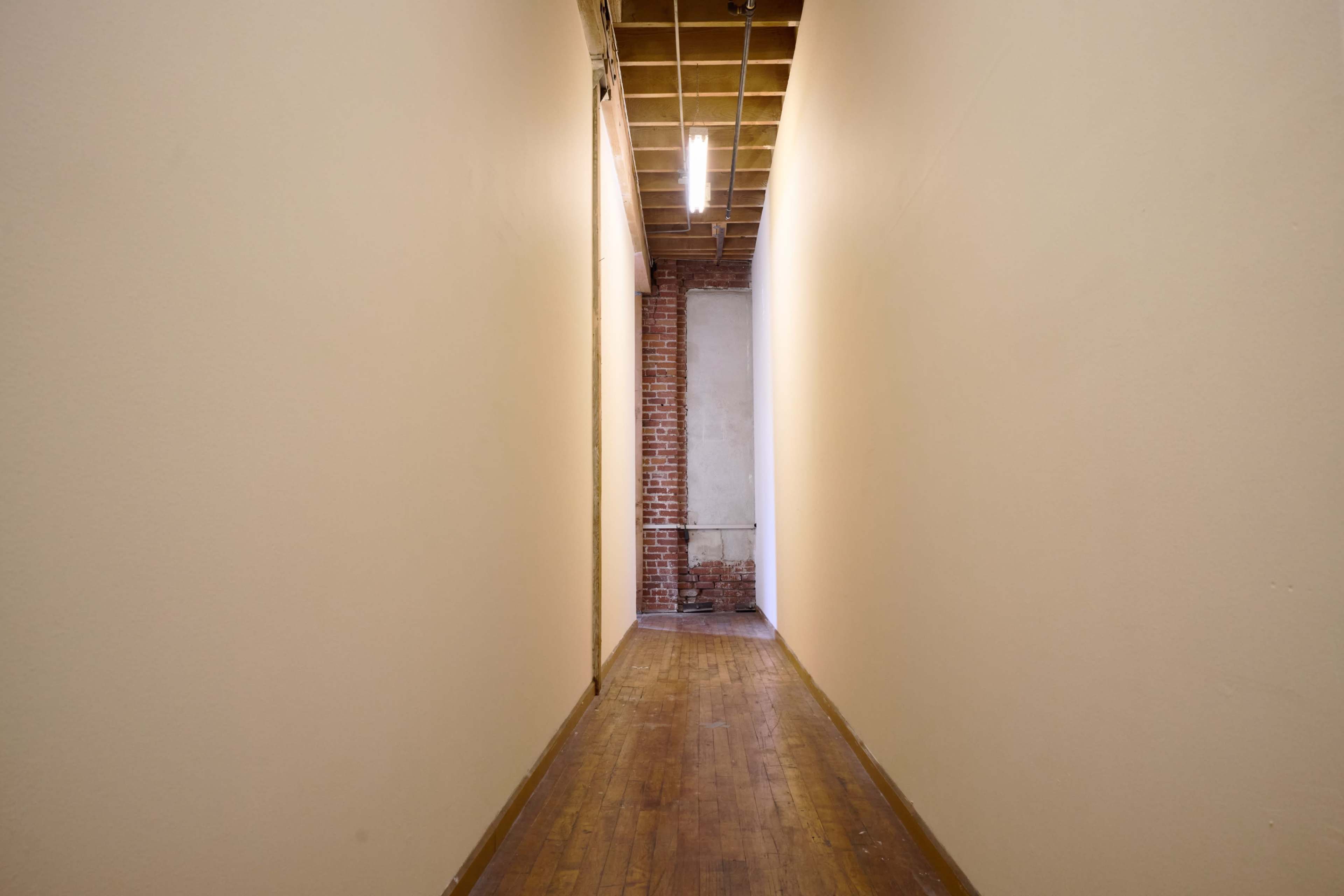 A narrow hallway with wooden flooring and light-colored walls extends towards a brick wall at the end.
