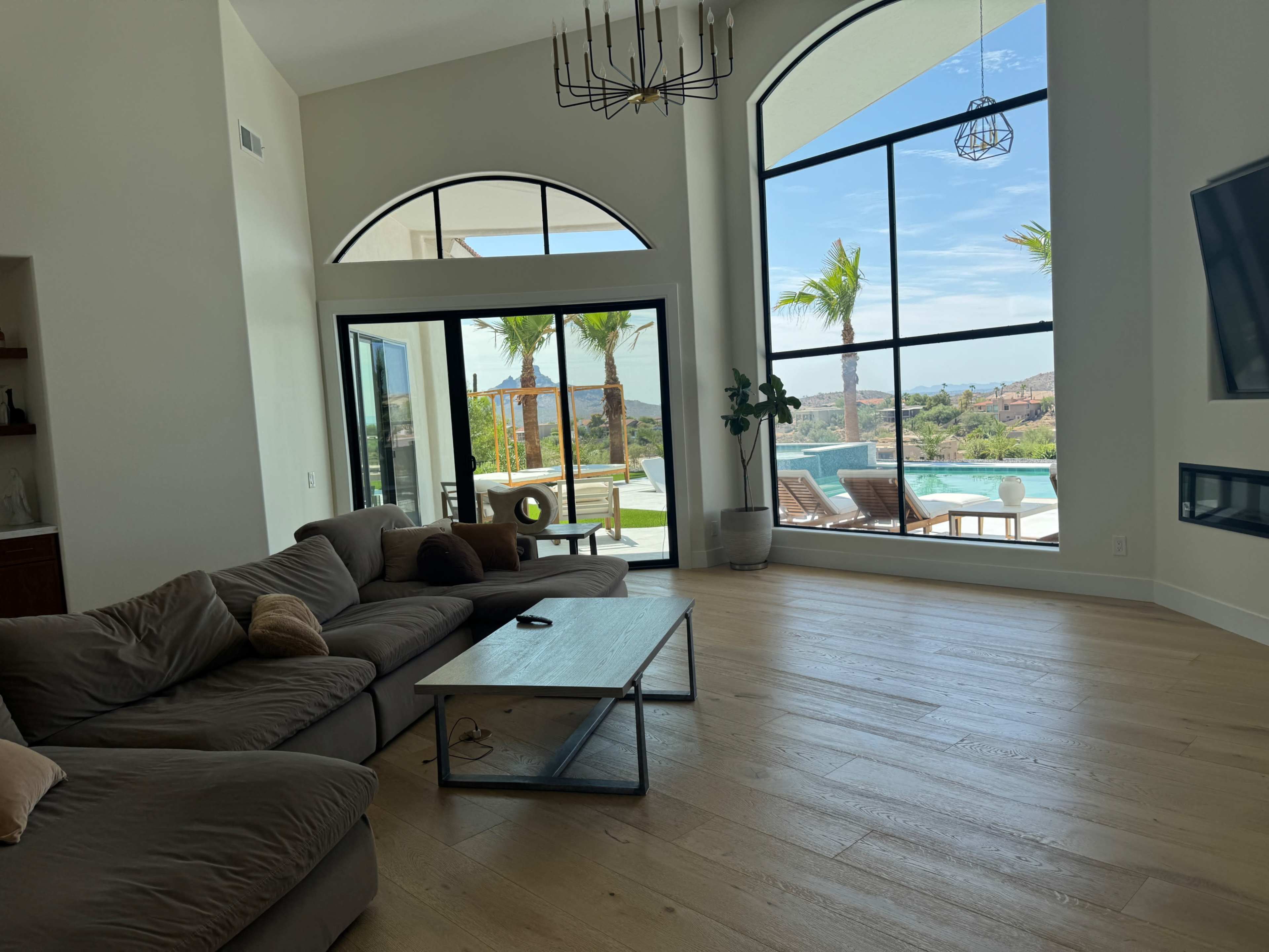 A spacious living room with a large sectional couch and a coffee table overlooks a pool area through tall glass doors framed by palm trees.