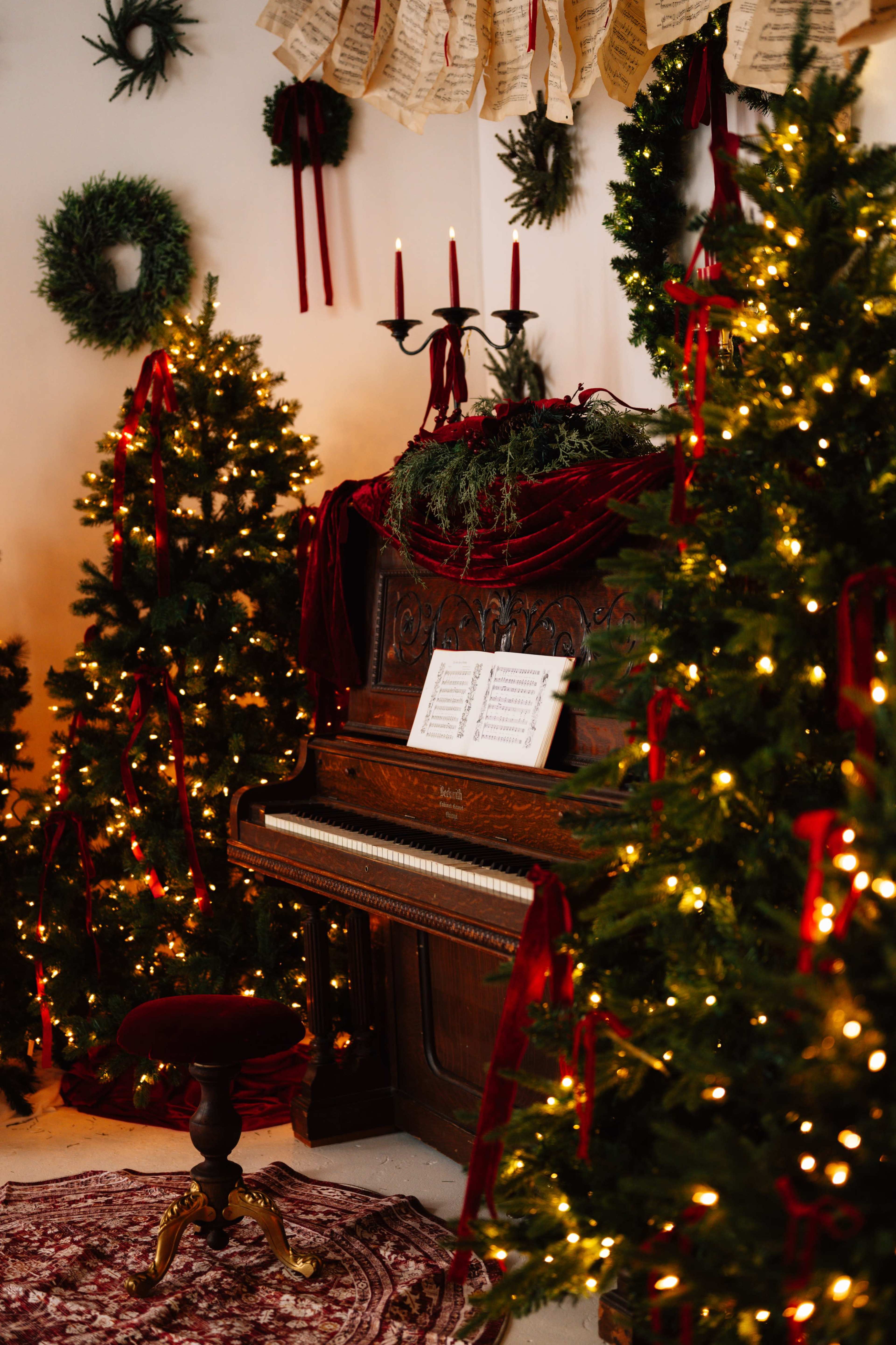 A vintage piano adorned with sheet music is surrounded by festive Christmas trees decorated with lights and ribbons in a cozy, holiday-themed room.