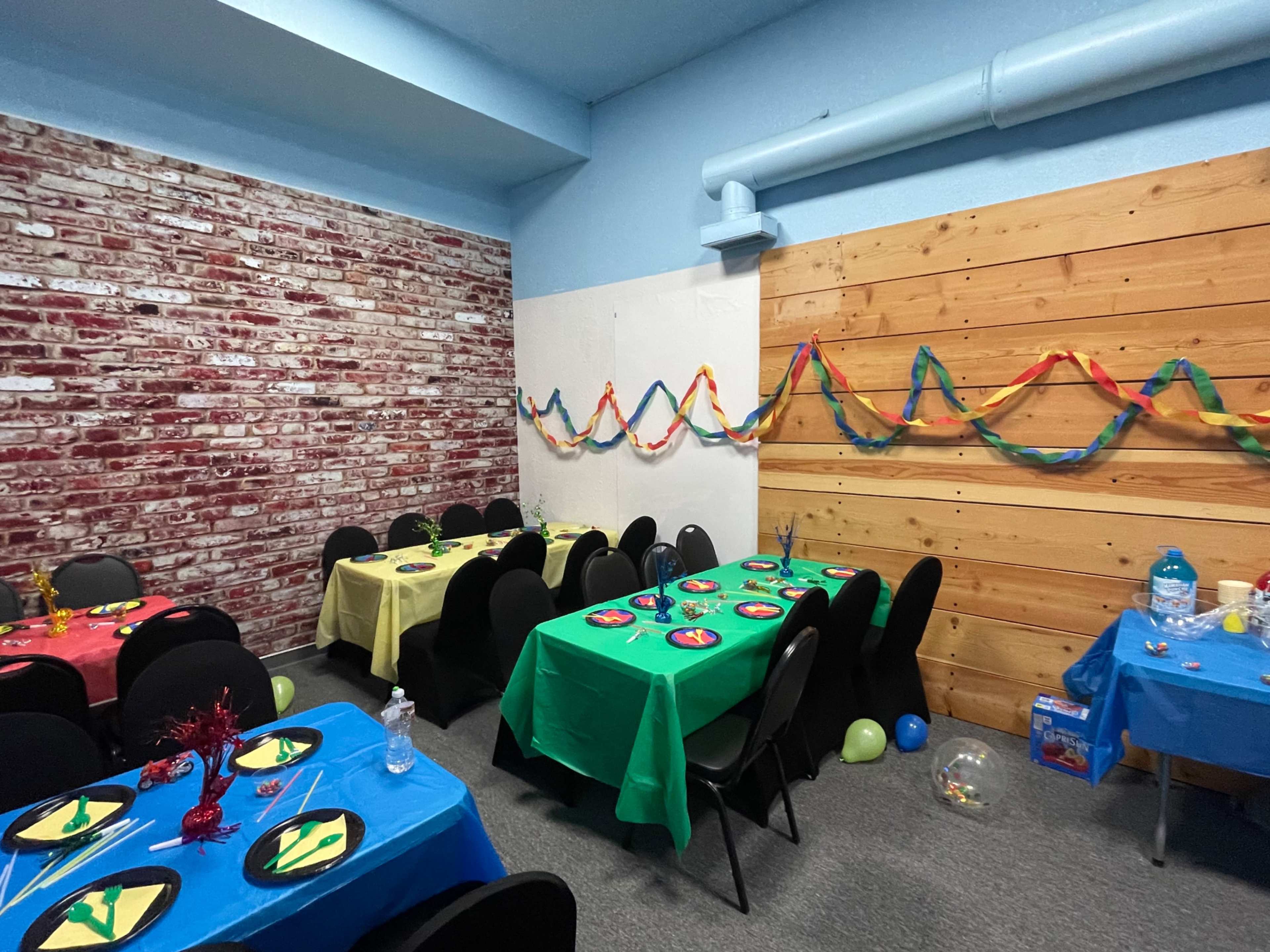 The image shows a decorated room with tables covered in bright tablecloths, featuring colorful decorations and party supplies against a backdrop of a brick wall and wooden paneling.