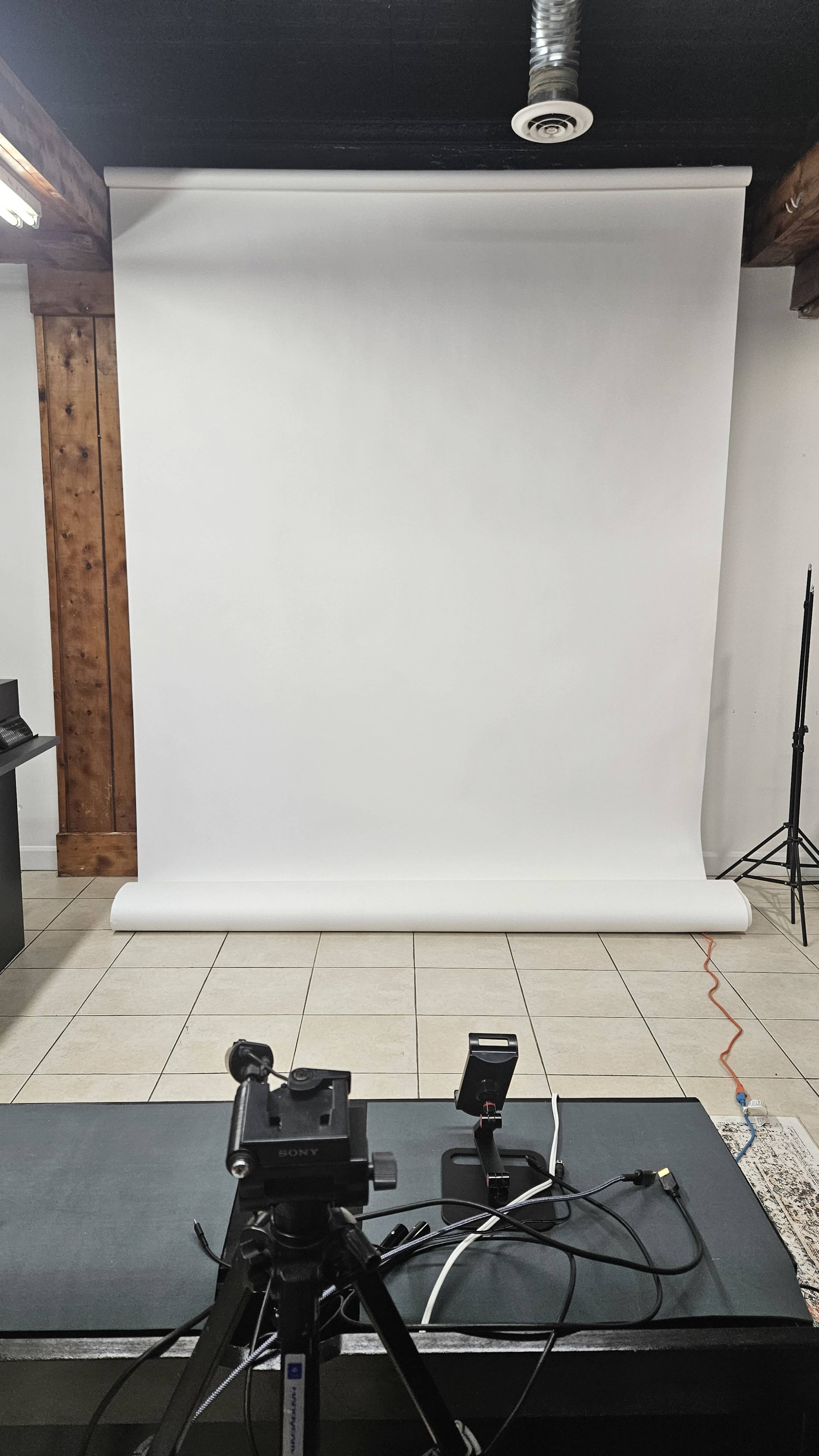 The image shows a photography studio with a white backdrop, a camera setup on a tripod, and various lighting equipment positioned nearby.