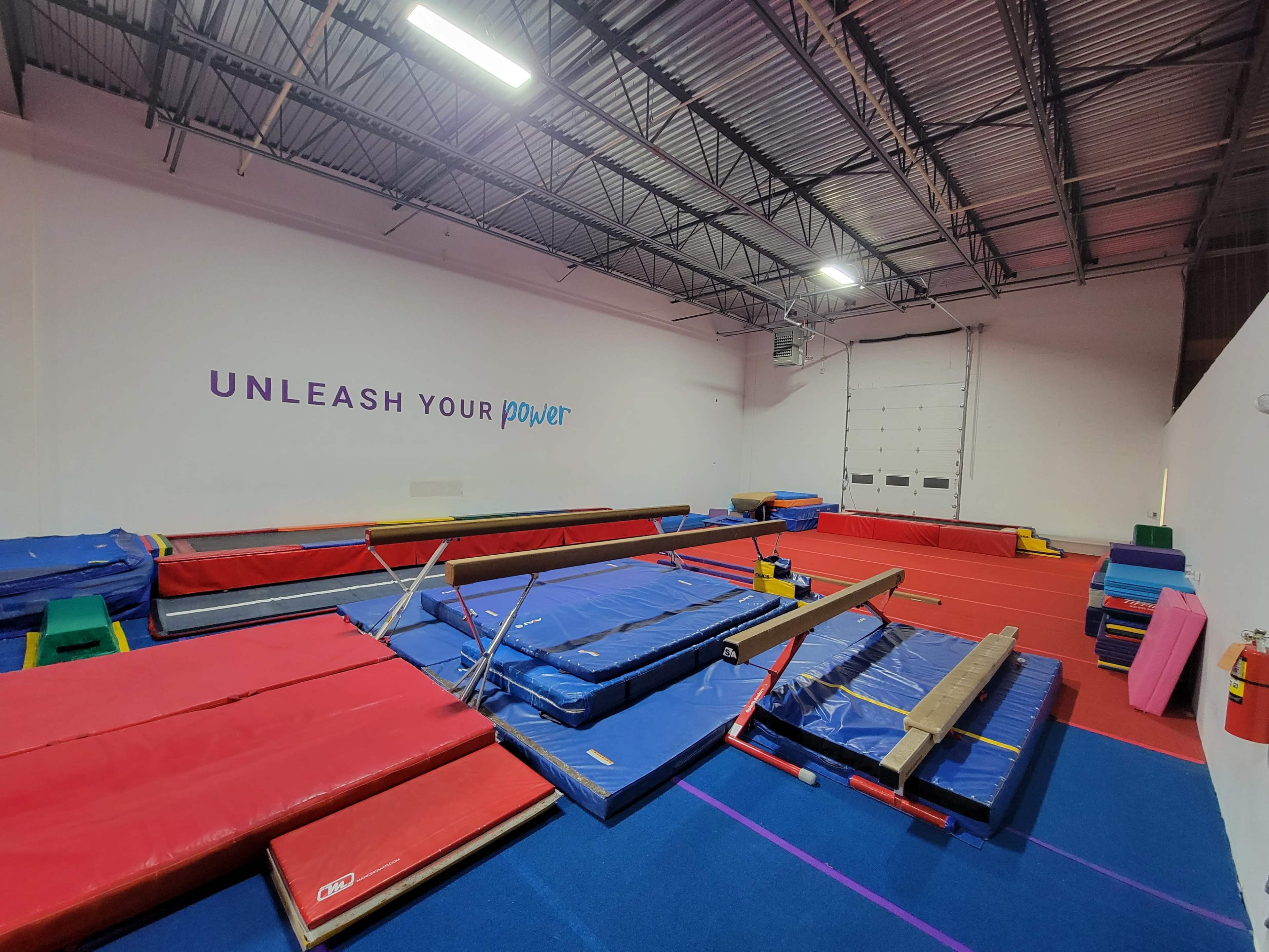 An indoor gymnastics facility with various mats, balance beams, and a wall featuring the text "UNLEASH YOUR power."