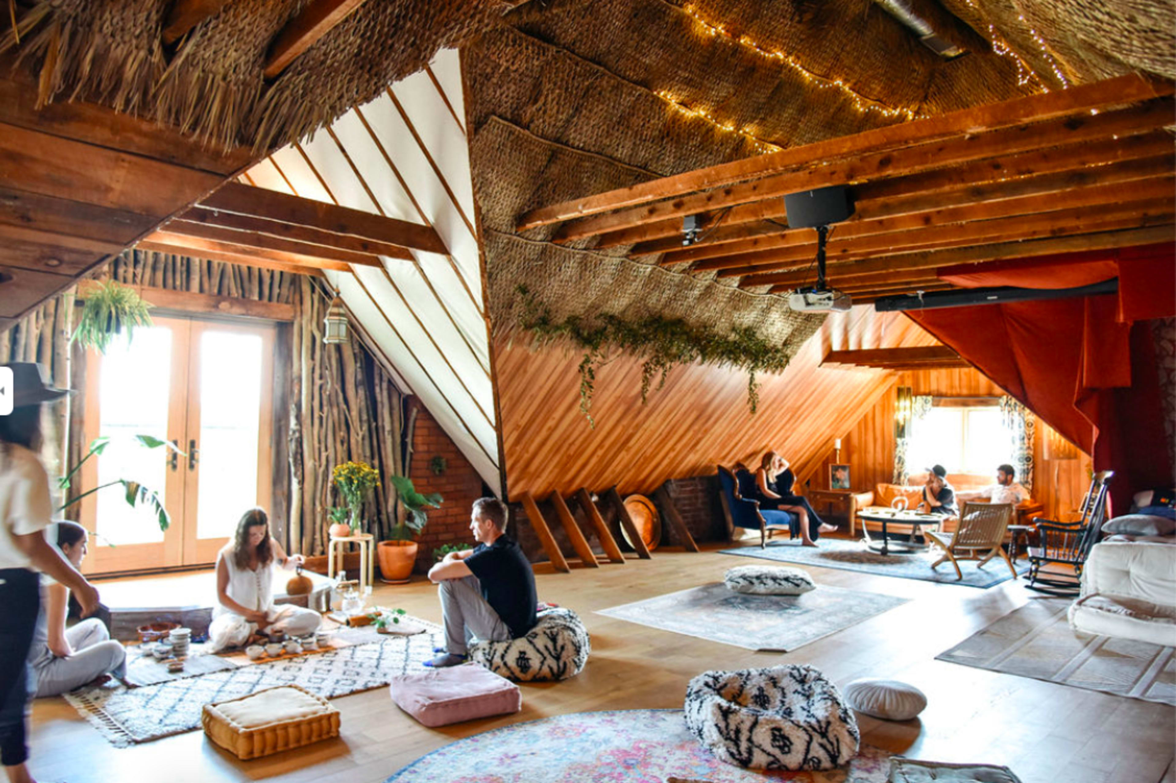 The scene features a spacious, wooden loft filled with people engaged in various activities, including lounging, socializing, and having tea, with decorative rugs and cushions scattered throughout.